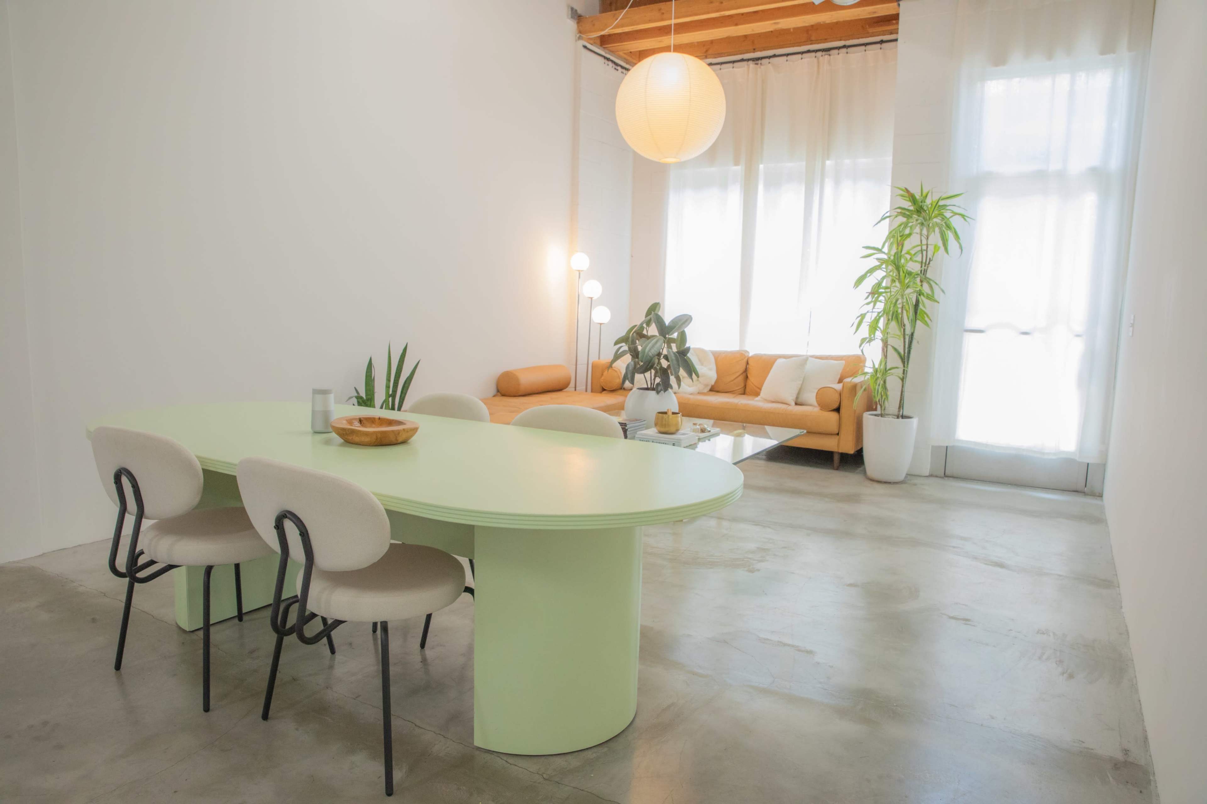 A light green oval dining table is set in a spacious room with modern furniture, including a brown sofa and plants near large windows.