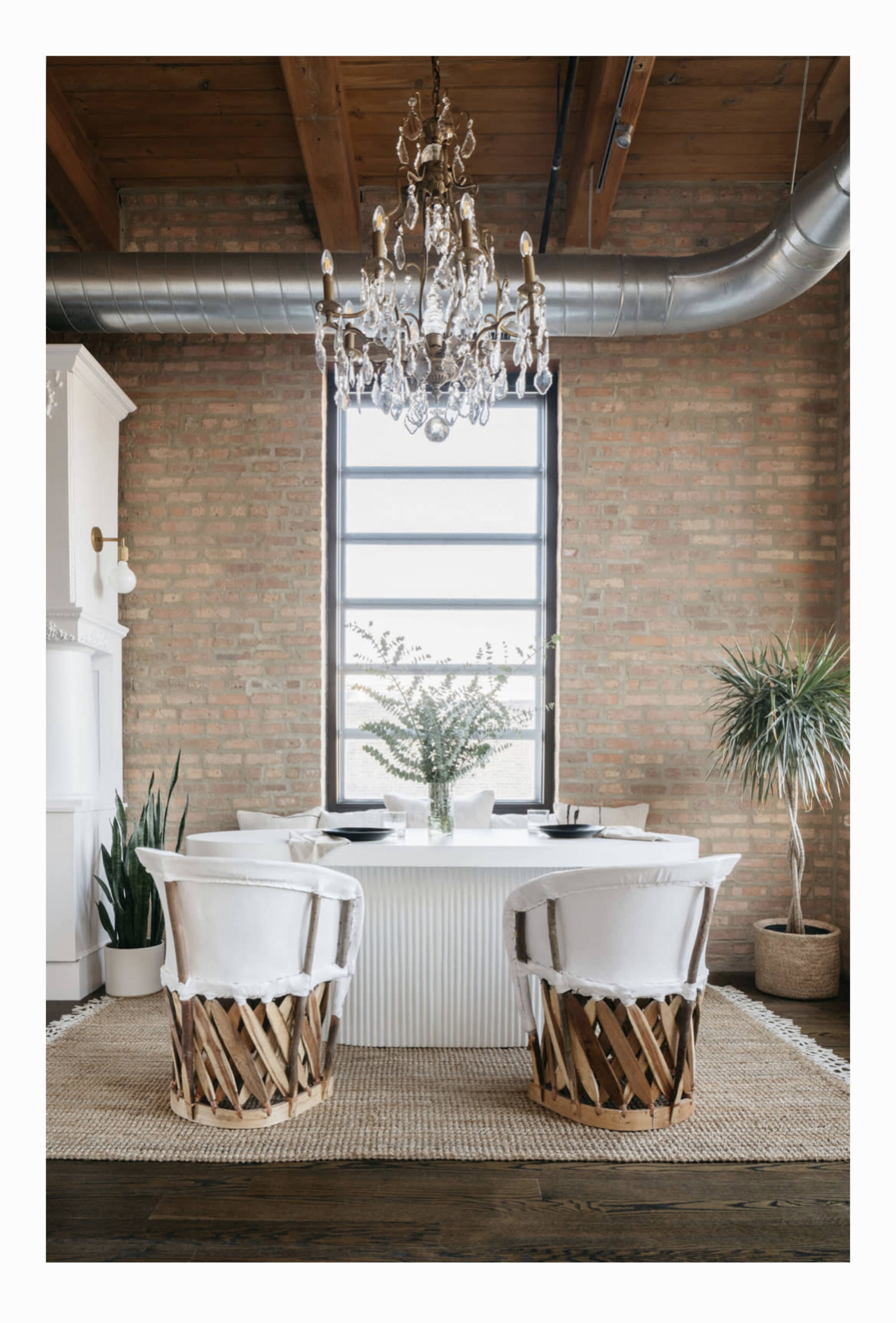 A dining area features a round table surrounded by two white chairs with wooden bases, alongside a large window and greenery.