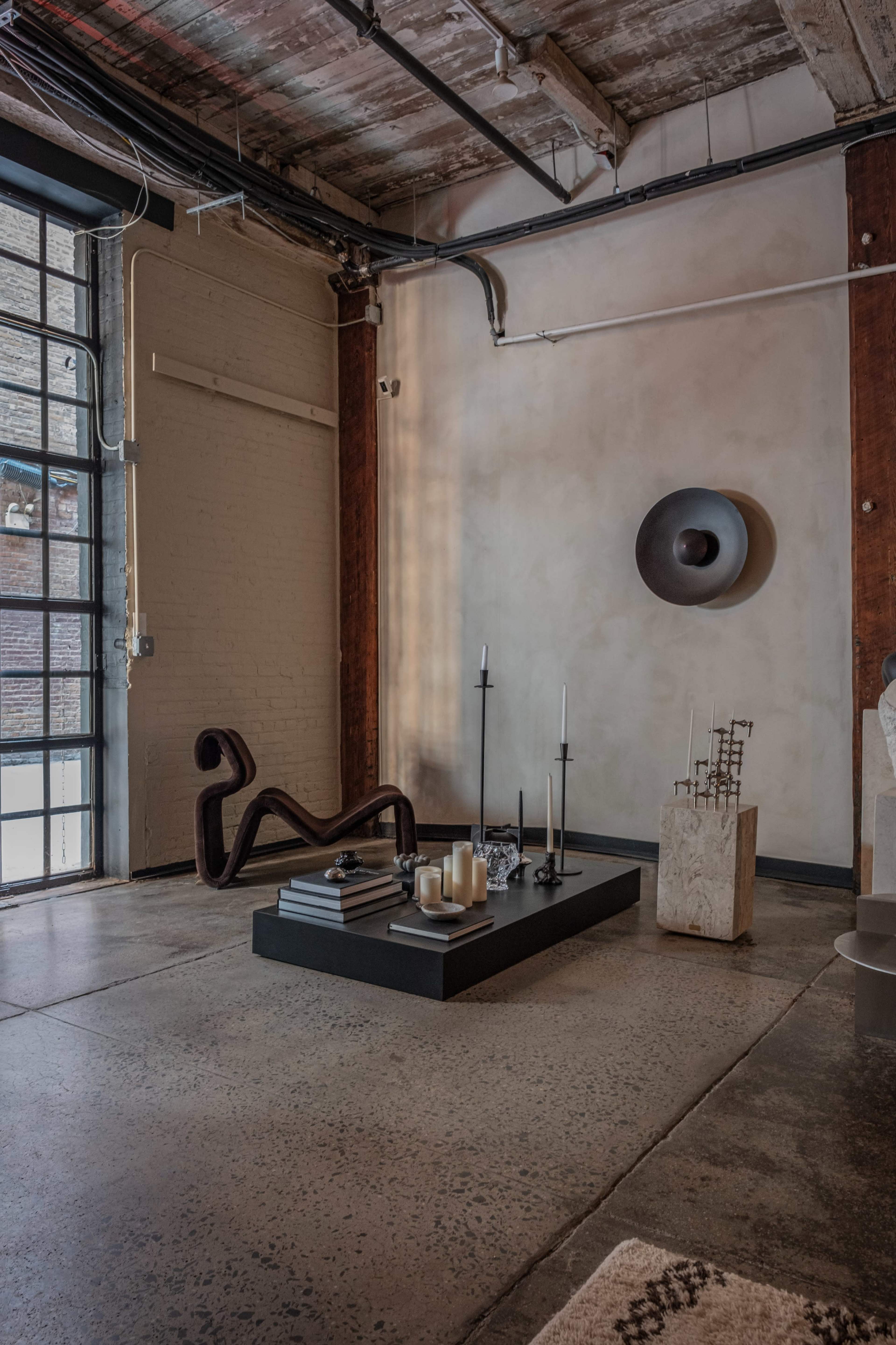 A minimalist interior space with a black platform featuring candles and sculptures, surrounded by large industrial windows and rustic walls.