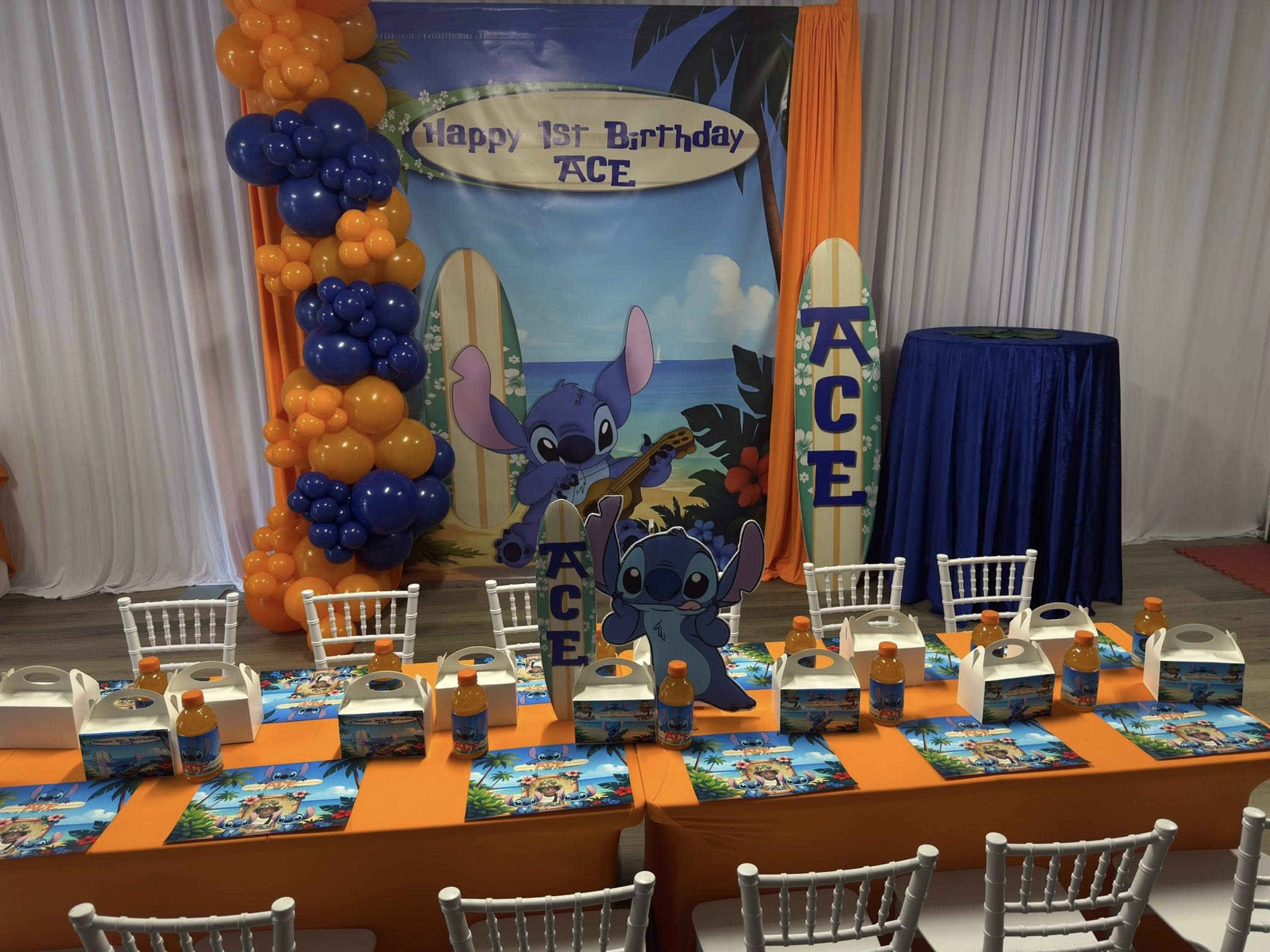 The scene features a brightly decorated birthday party setup for a one-year-old, with a tropical theme, complete with a backdrop, surfboards, and themed decorations on the tables.