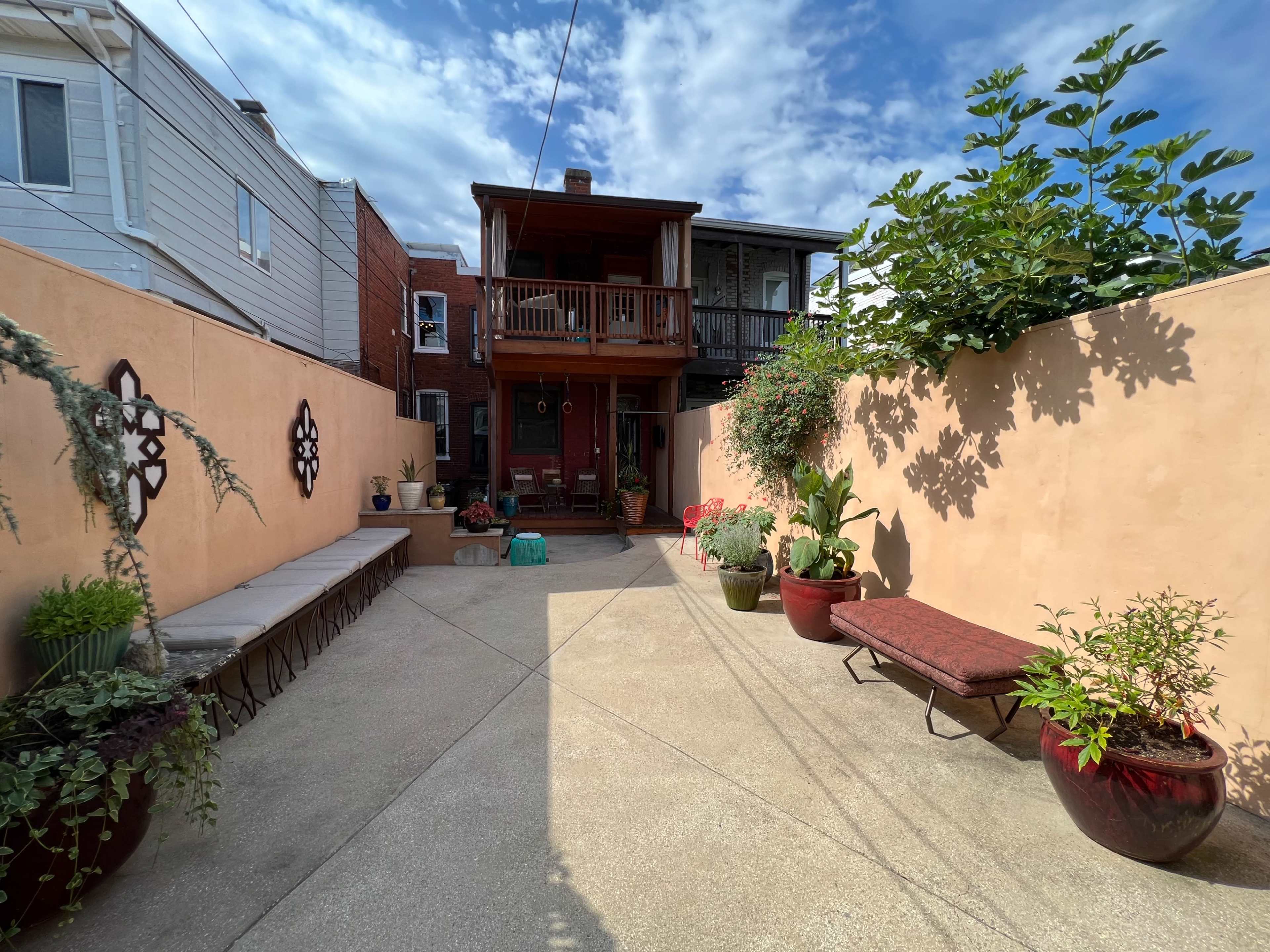The image shows a narrow courtyard with a concrete surface, bordered by orange walls, and featuring potted plants and a bench, leading to a wooden balcony in the background.