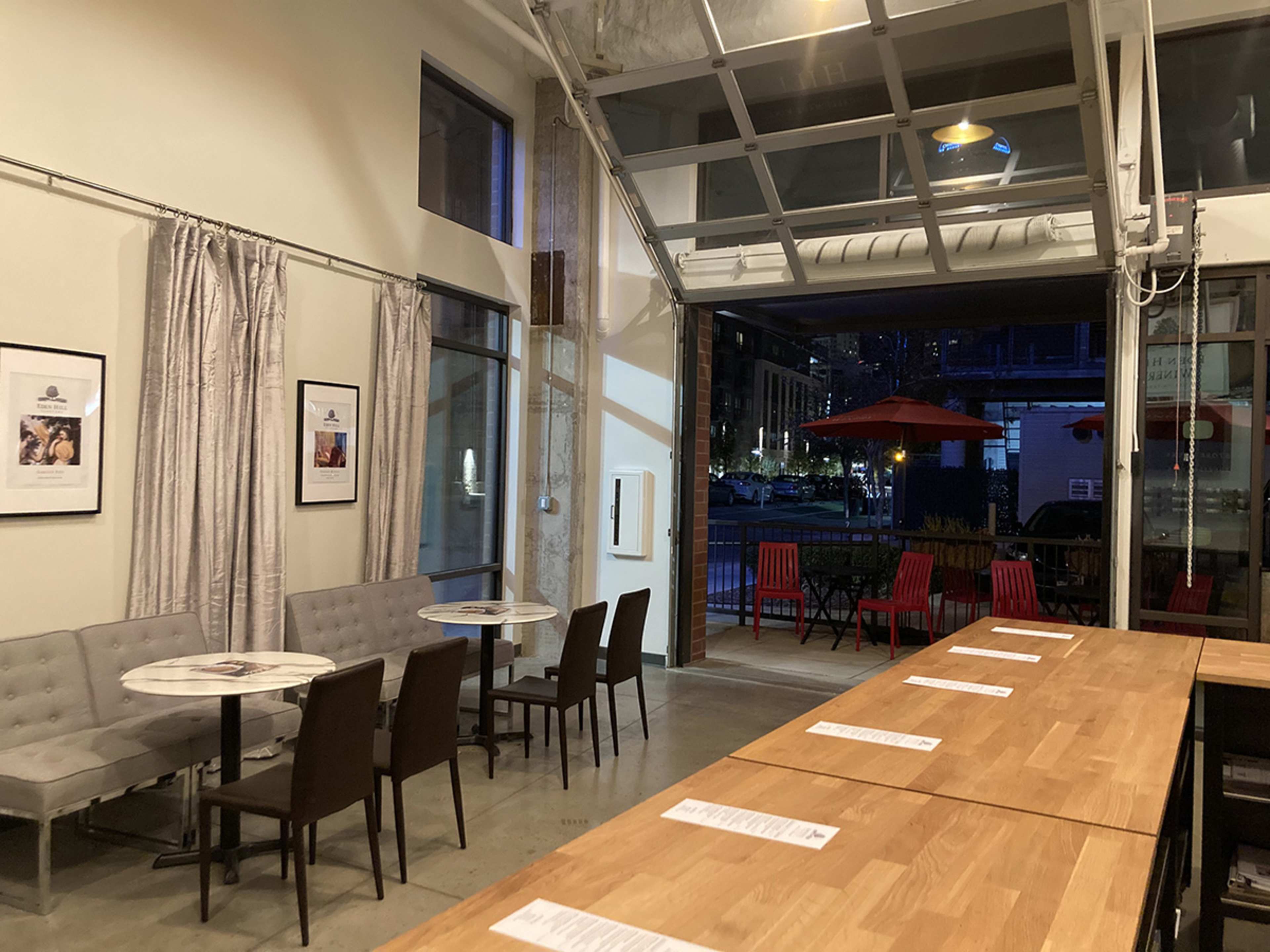 A modern café interior with tables and chairs arranged near an open garage door that leads to an outdoor seating area.