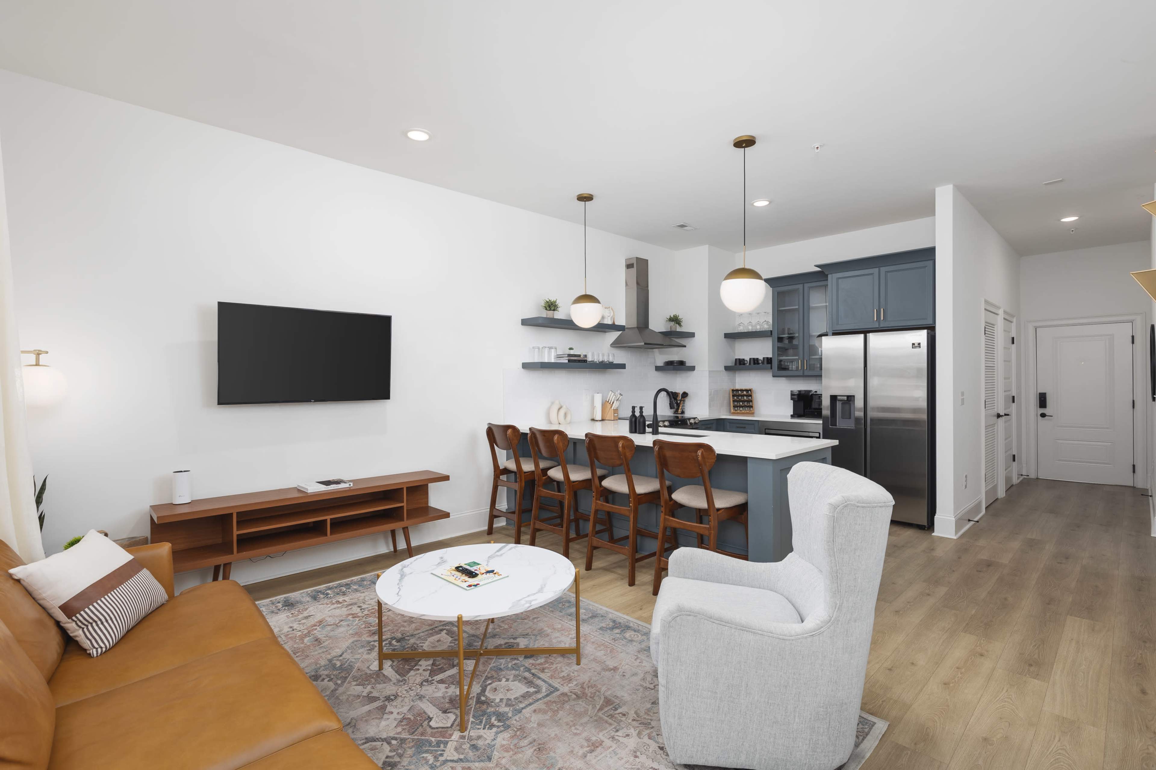 The image shows a modern living space featuring a leather sofa, a round coffee table, a dining area with wooden chairs, and a kitchen with stainless steel appliances.