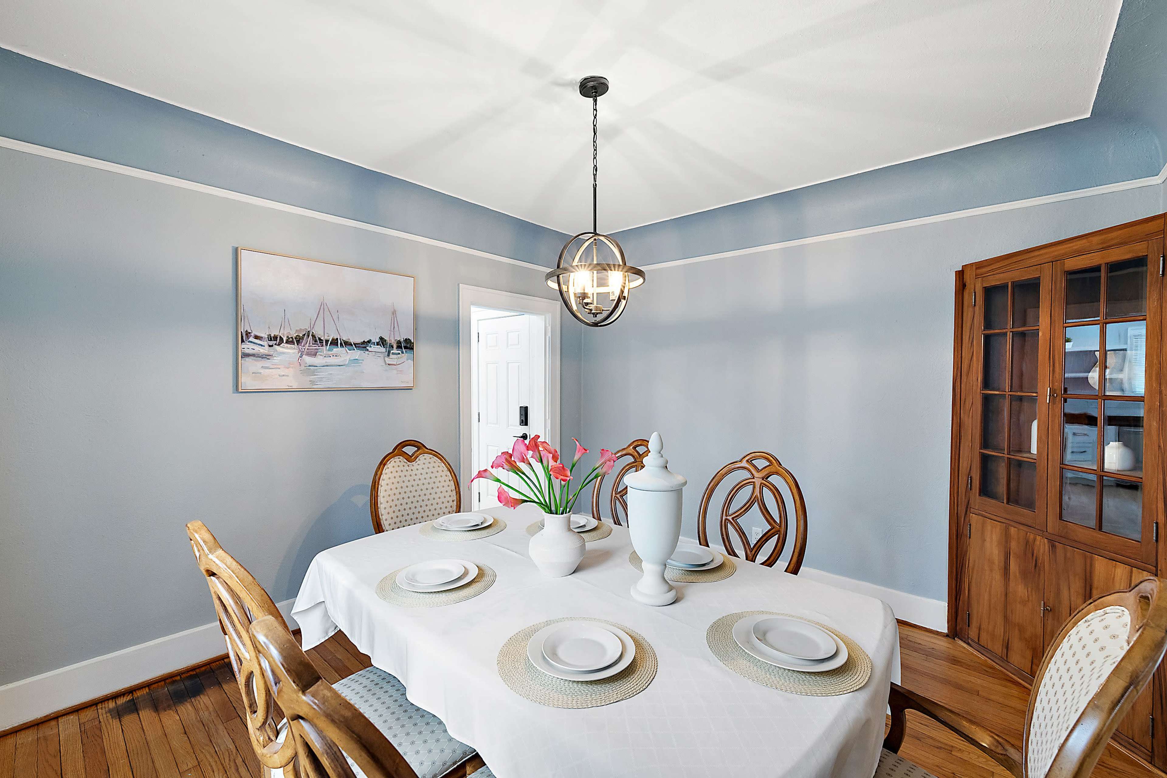A dining room features a round table set with plates and a vase of tulips, surrounded by wooden chairs, with a cabinet and a painting on the walls.