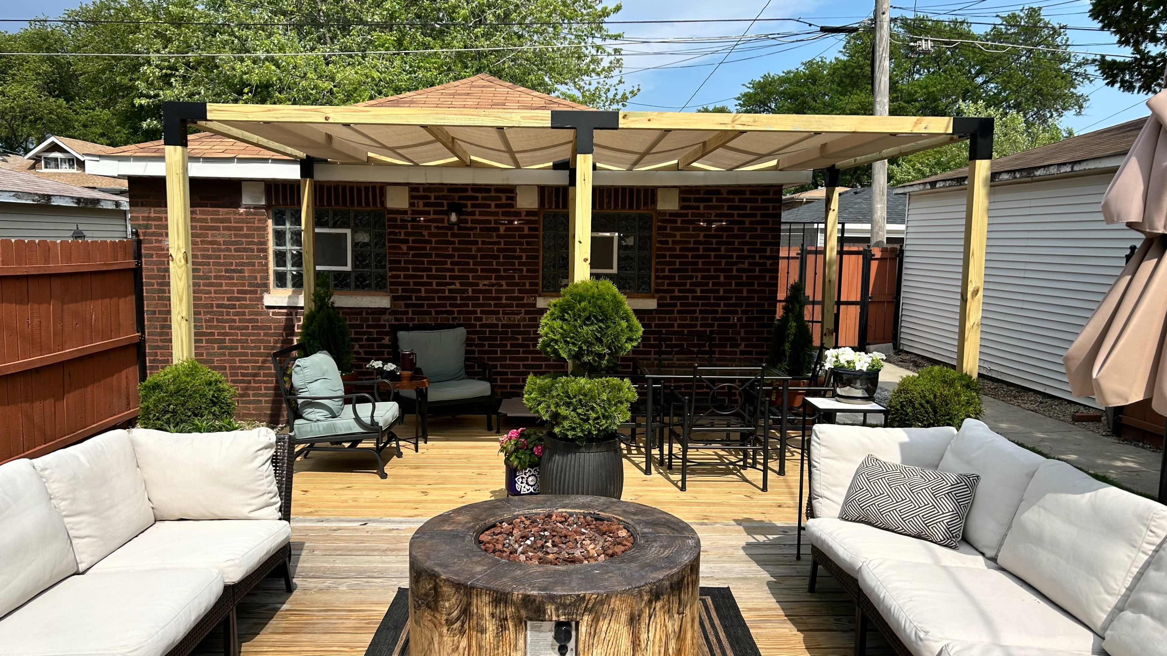 The image shows a backyard patio area with a wooden deck, featuring a fire pit surrounded by seating, a pergola overhead, and landscaping with potted plants.
