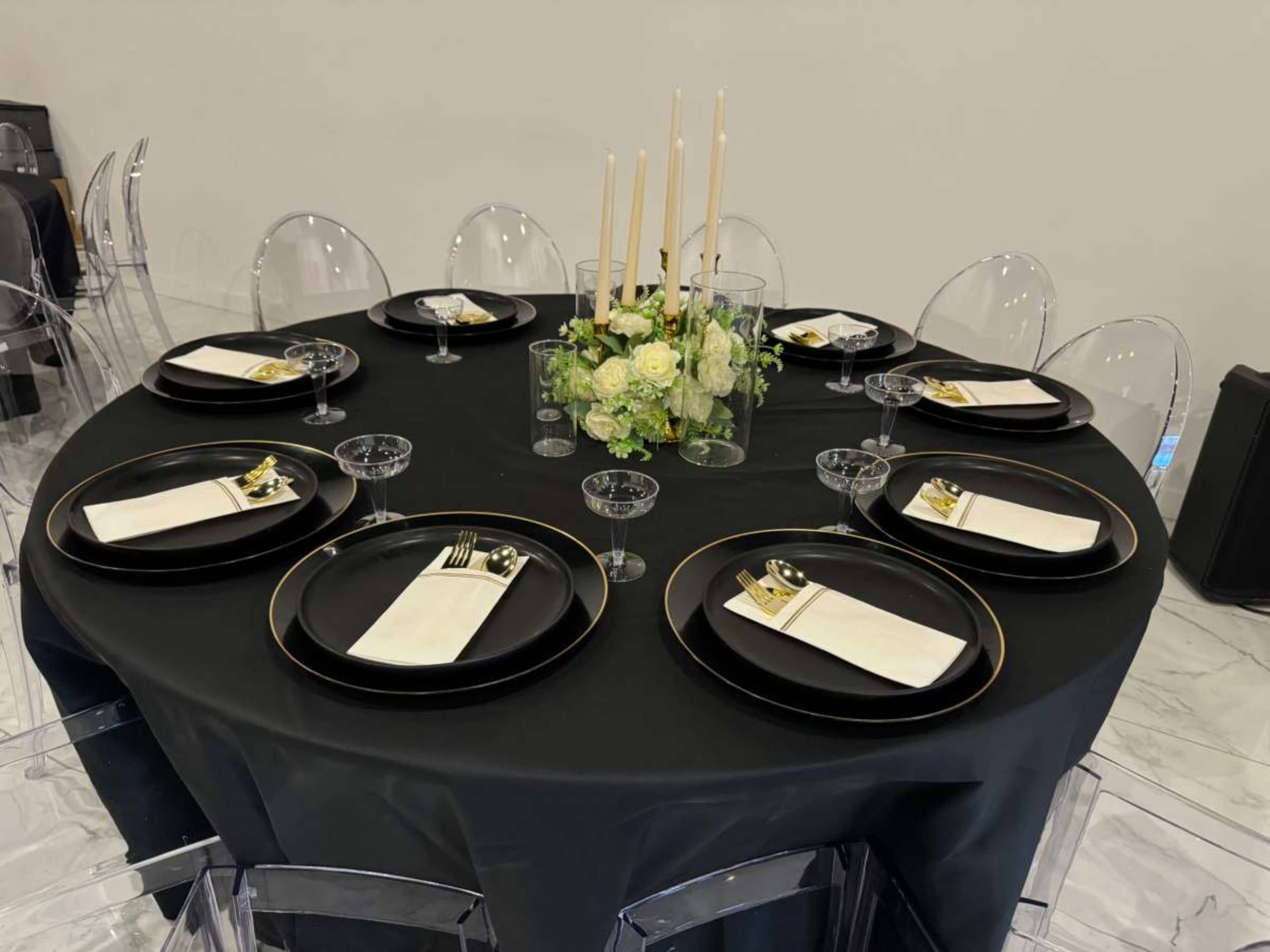 A round table is set with black plates, white napkins, glassware, and a floral centerpiece featuring candles.