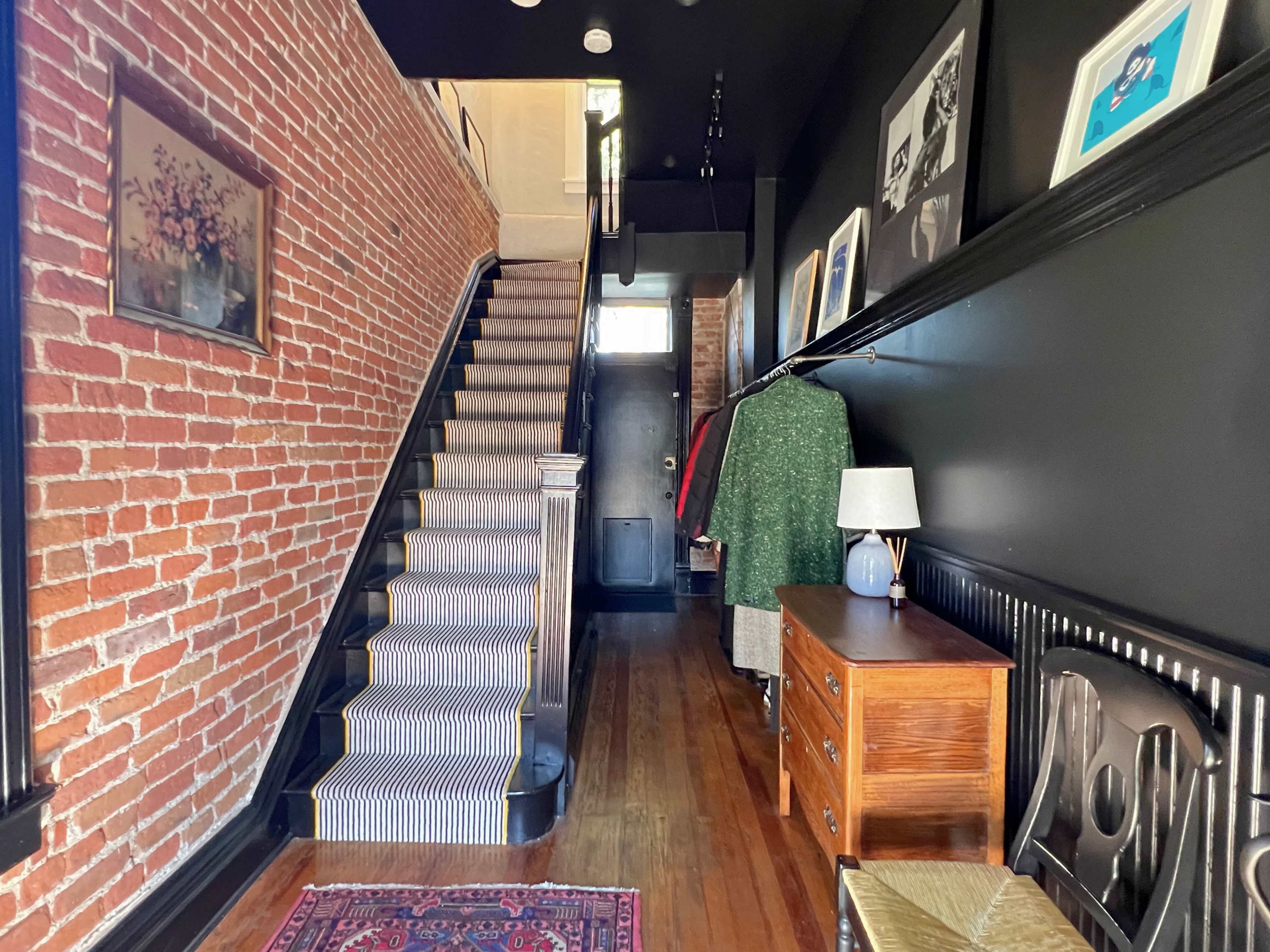 The image shows a hallway with exposed brick walls, stairs leading to an upper floor, and a decorative wooden dresser against the wall.