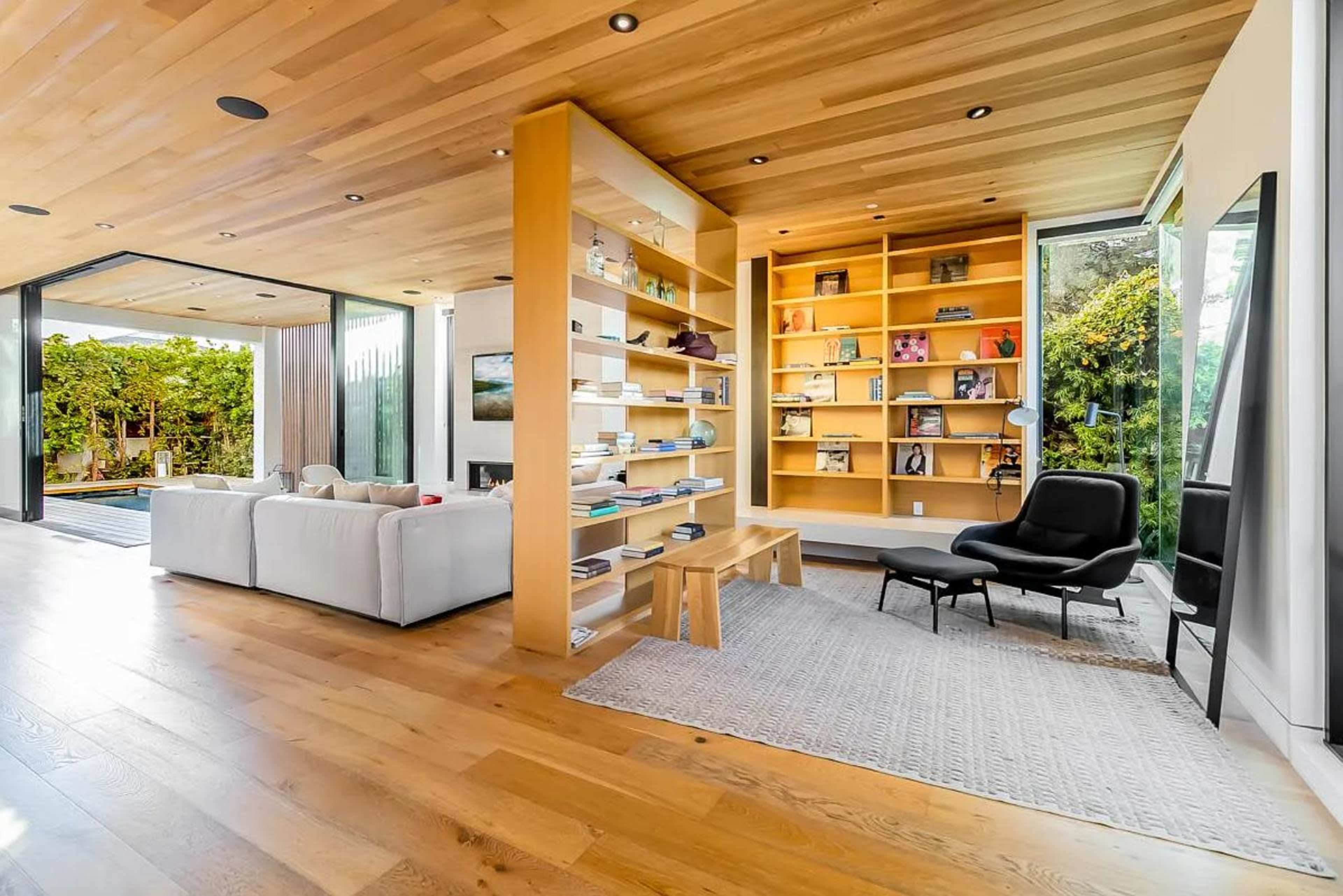 The image shows a modern living space featuring a wooden ceiling, a bookshelf filled with items, a comfortable black chair, and large windows that provide a view of greenery outside.