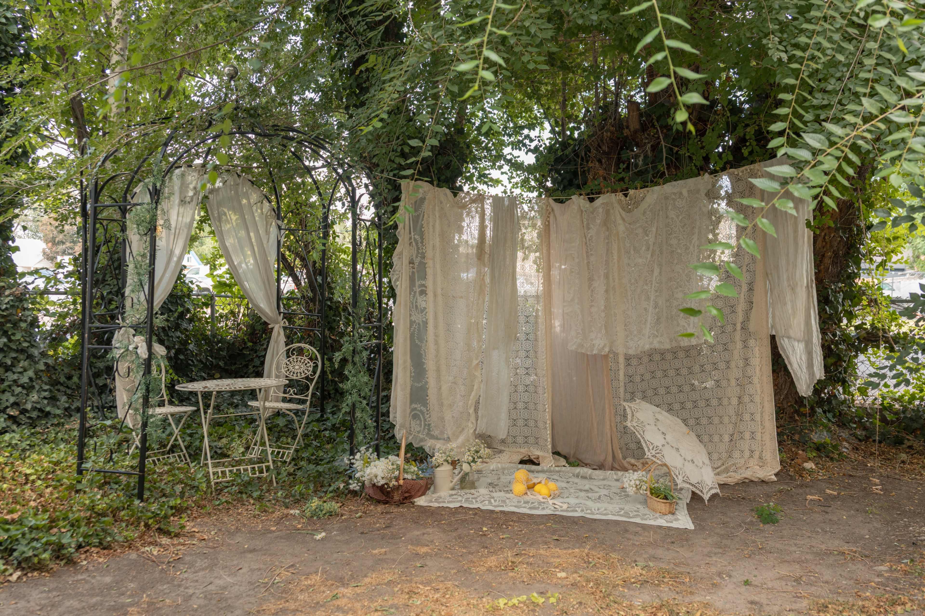 A decorative outdoor space featuring a wrought iron archway with a small table and chairs, draped with lace curtains and surrounded by greenery.