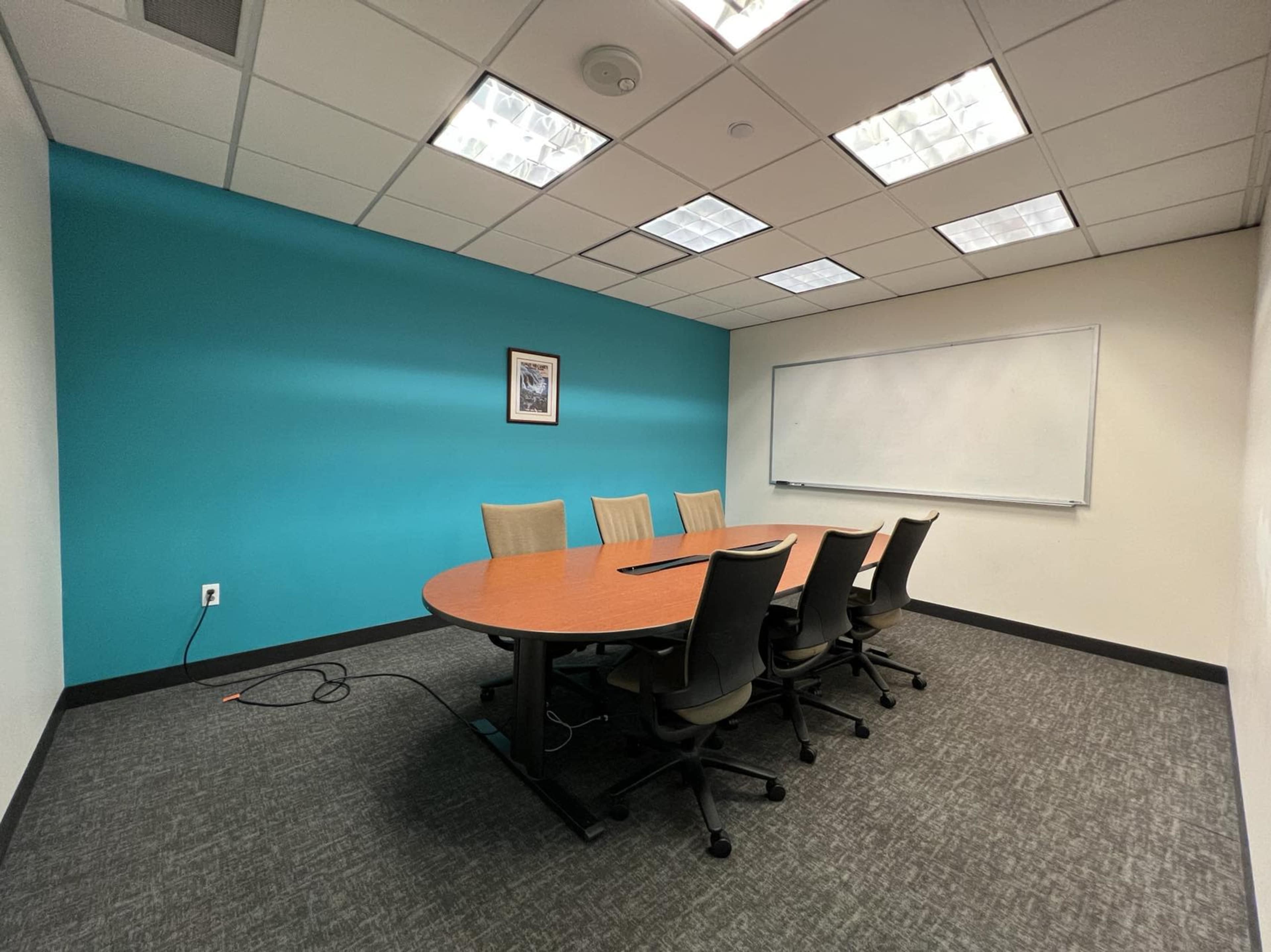 The image shows a meeting room featuring a large round table with six office chairs, a whiteboard, and a blue accent wall.