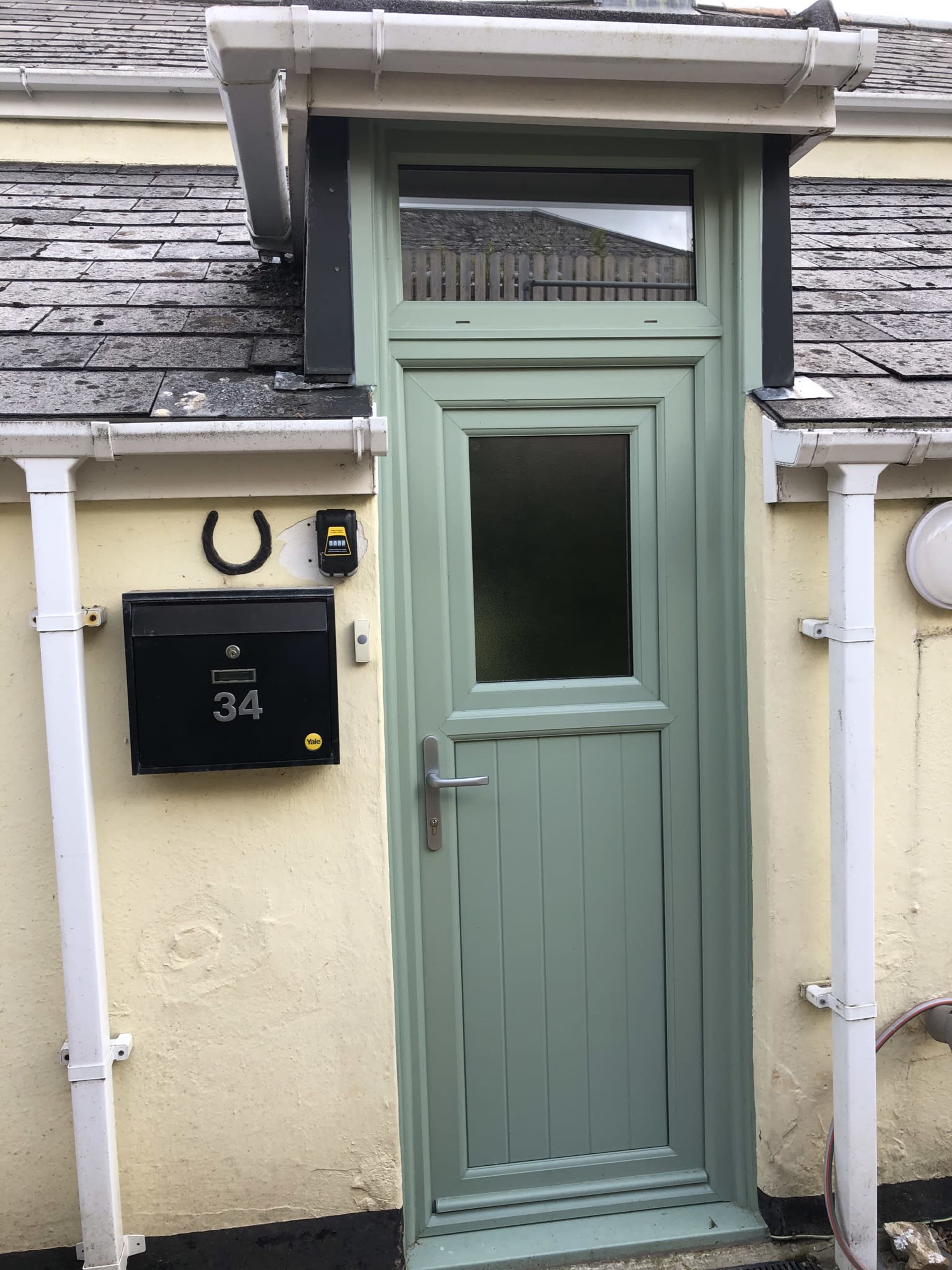 A green door with a small window above it, next to a black mailbox displaying the number 34, against a yellow wall.