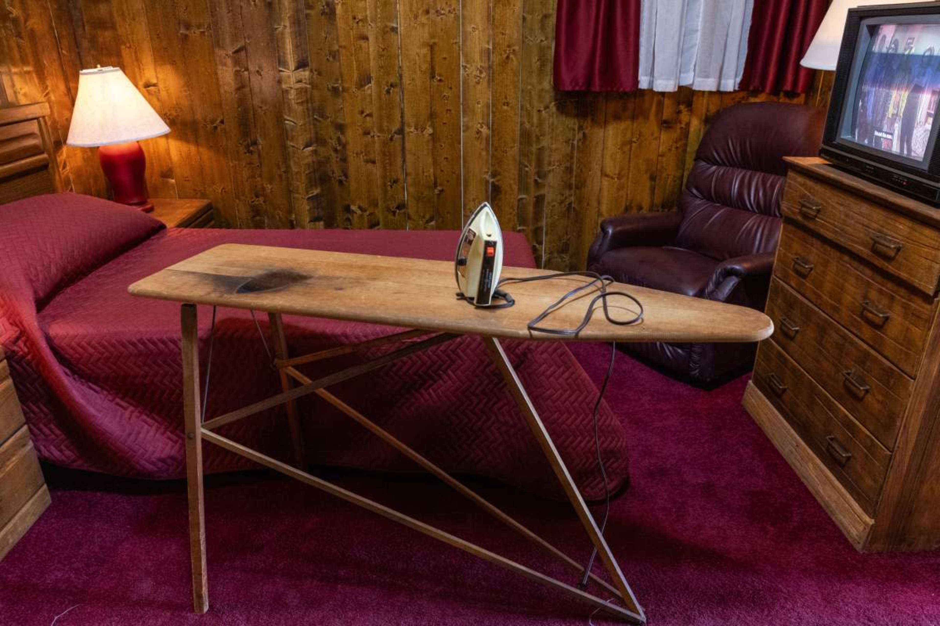 The image shows a simple bedroom with a bed, an ironing board with an iron on it, a television, and a recliner chair.