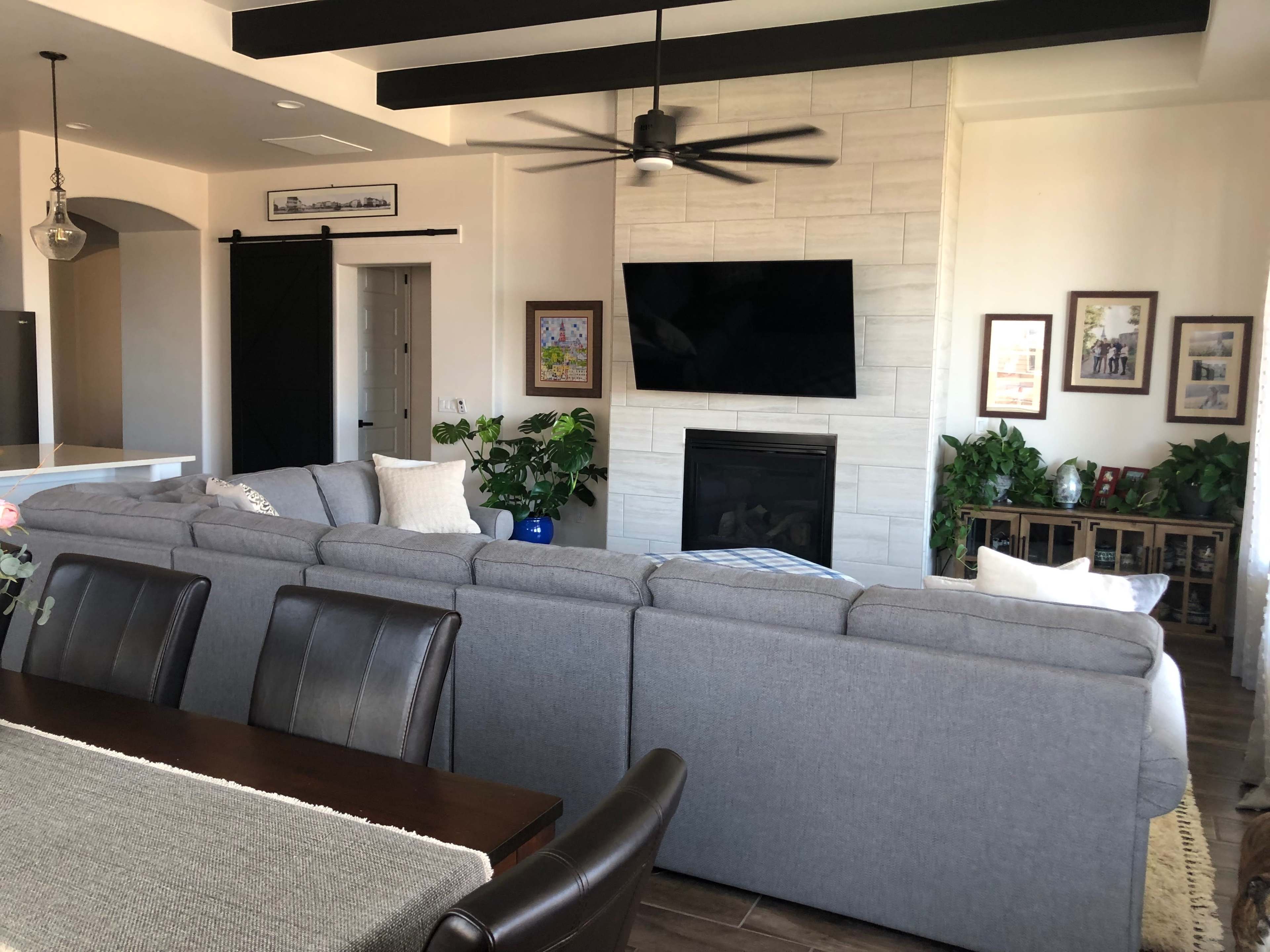 The image shows a living room with a large gray sectional sofa, a mounted television above a fireplace, and various indoor plants arranged throughout the space.