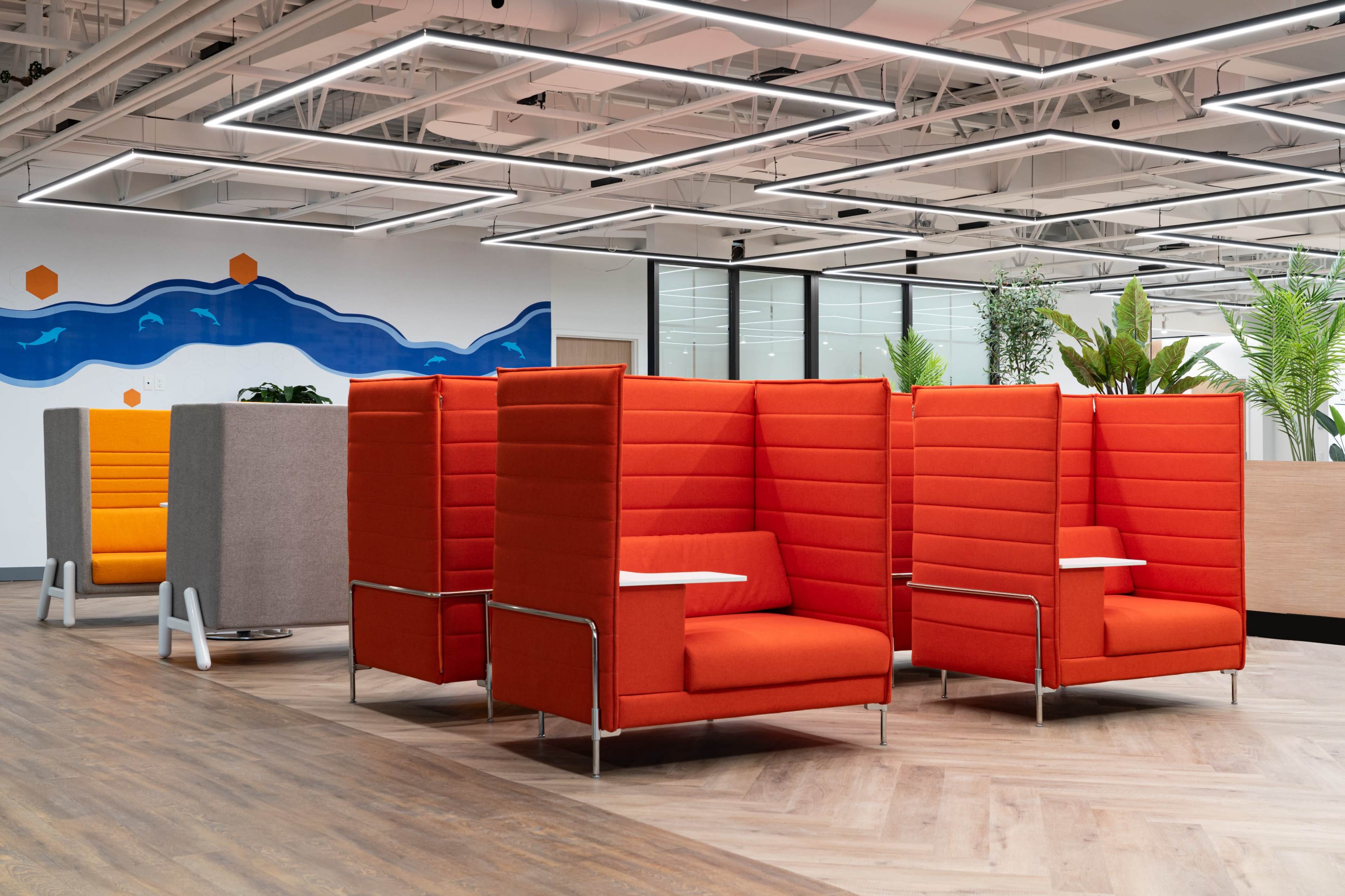 A modern office space featuring orange and gray seating pods with attached side tables, surrounded by indoor plants and decorative wall art.