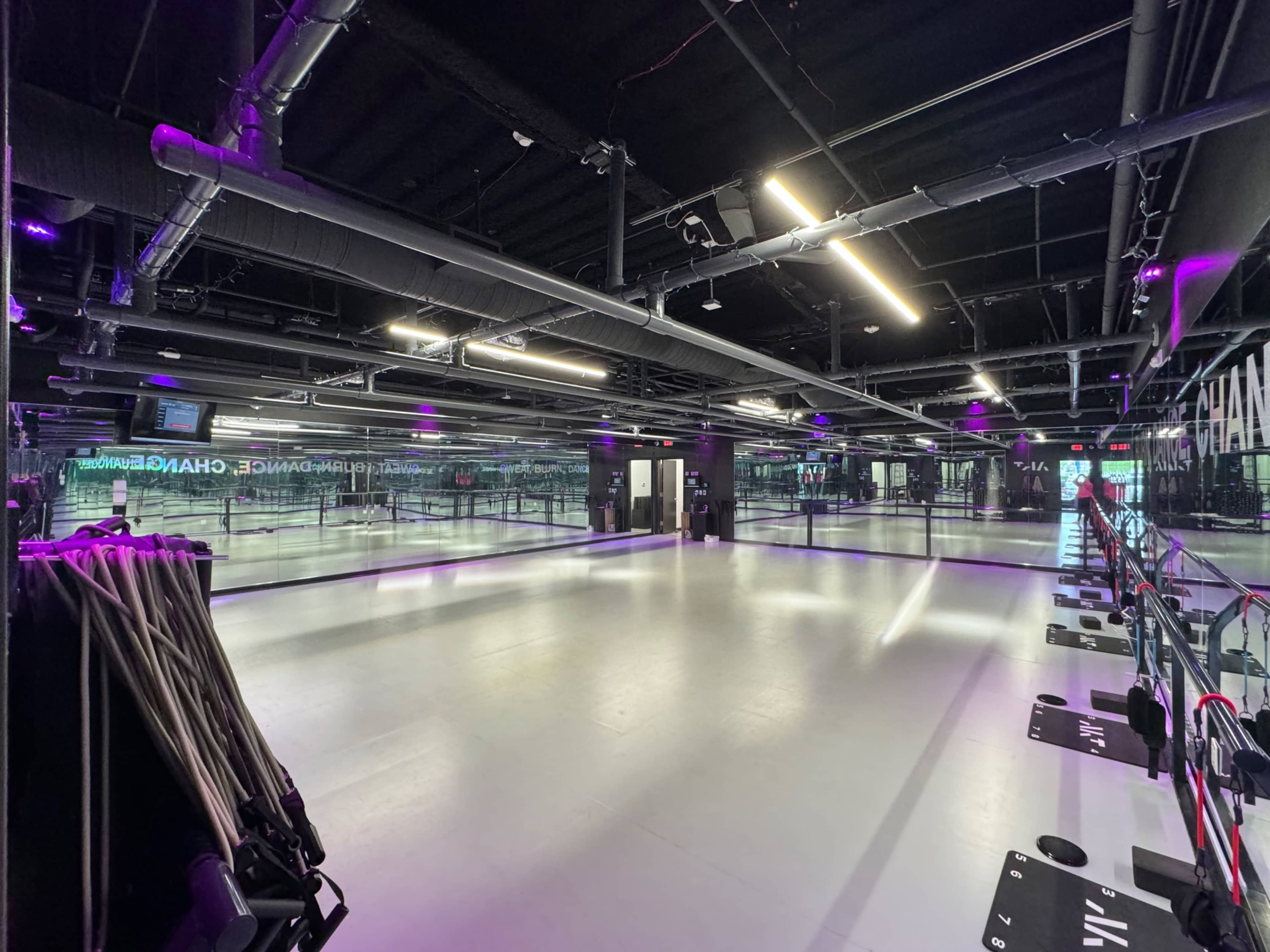 A spacious gym interior with black walls, purple lighting, and exercise equipment lined up along one side.