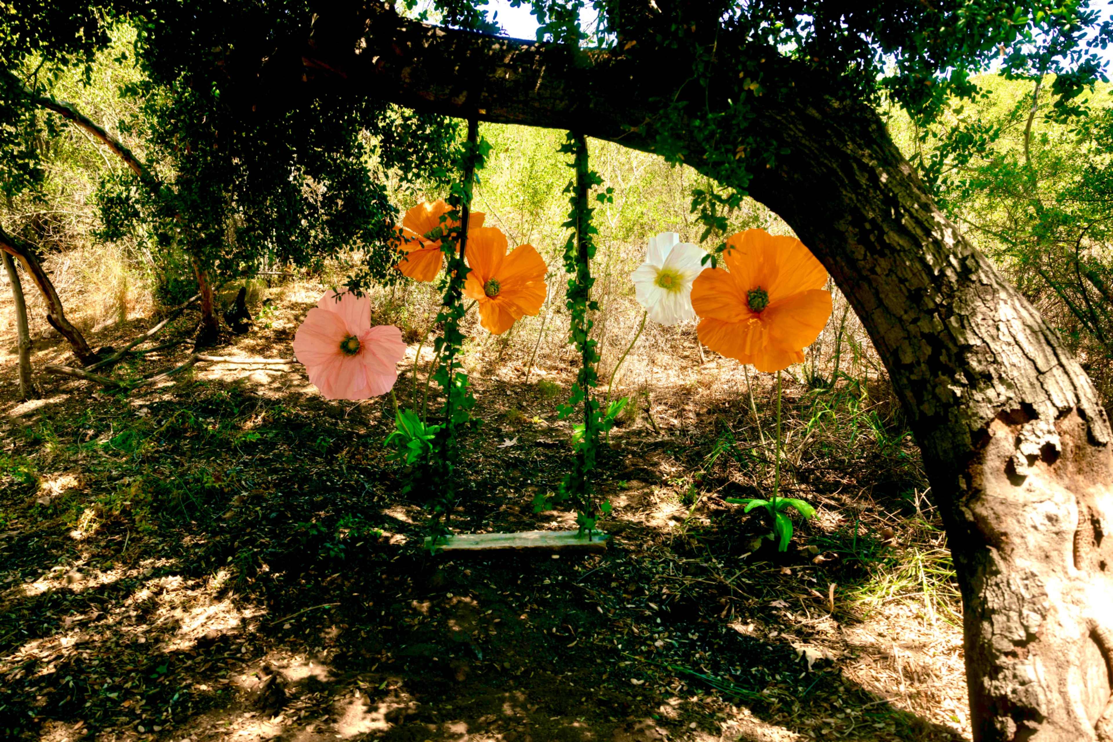 A swing adorned with large, colorful paper flowers hangs from a tree in a wooded area.