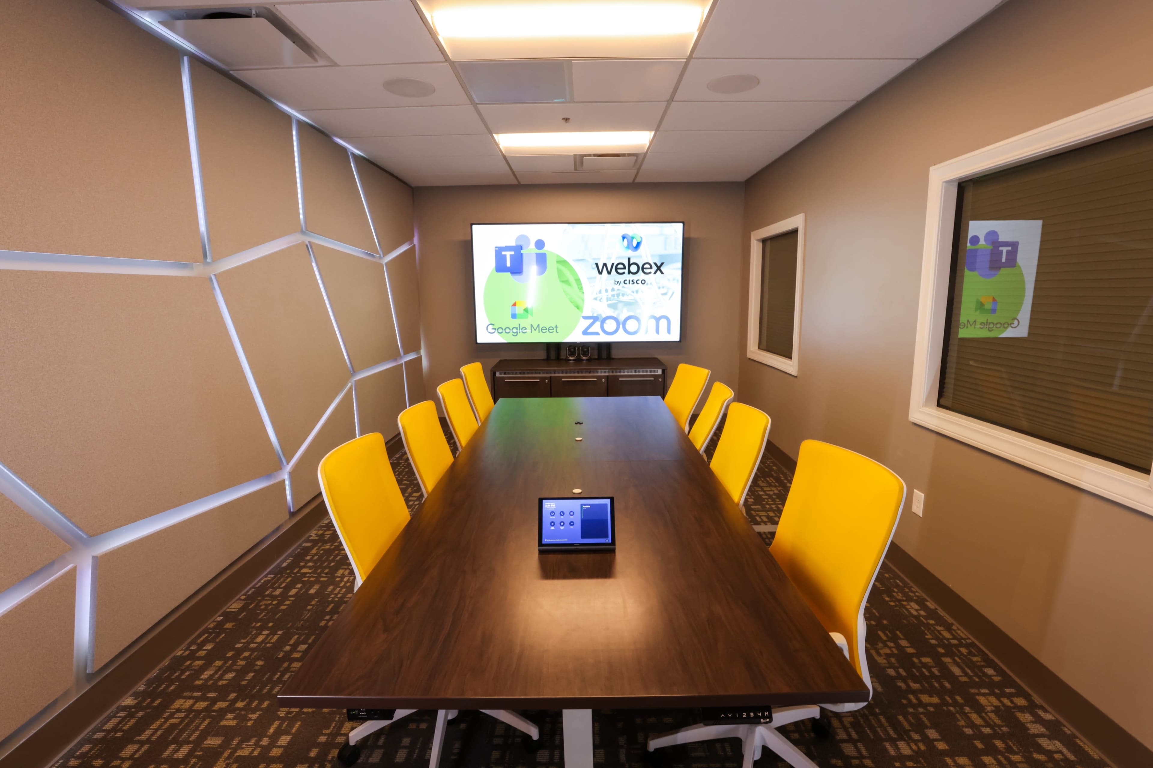 A modern conference room features a large table surrounded by yellow chairs, with a screen displaying multiple video conferencing platform logos.