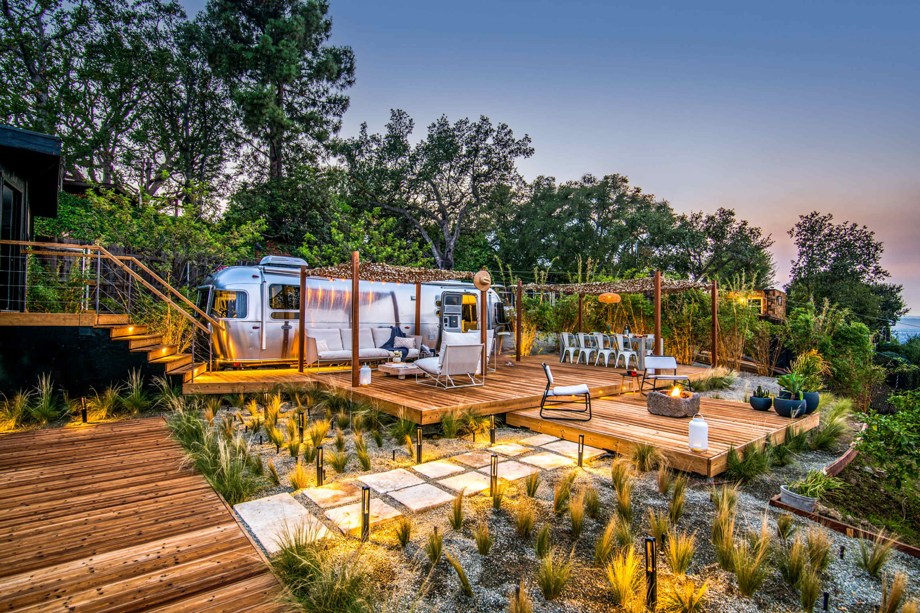 A modern outdoor space features a vintage Airstream trailer, surrounded by wooden decking, seating areas, and landscaped greenery.