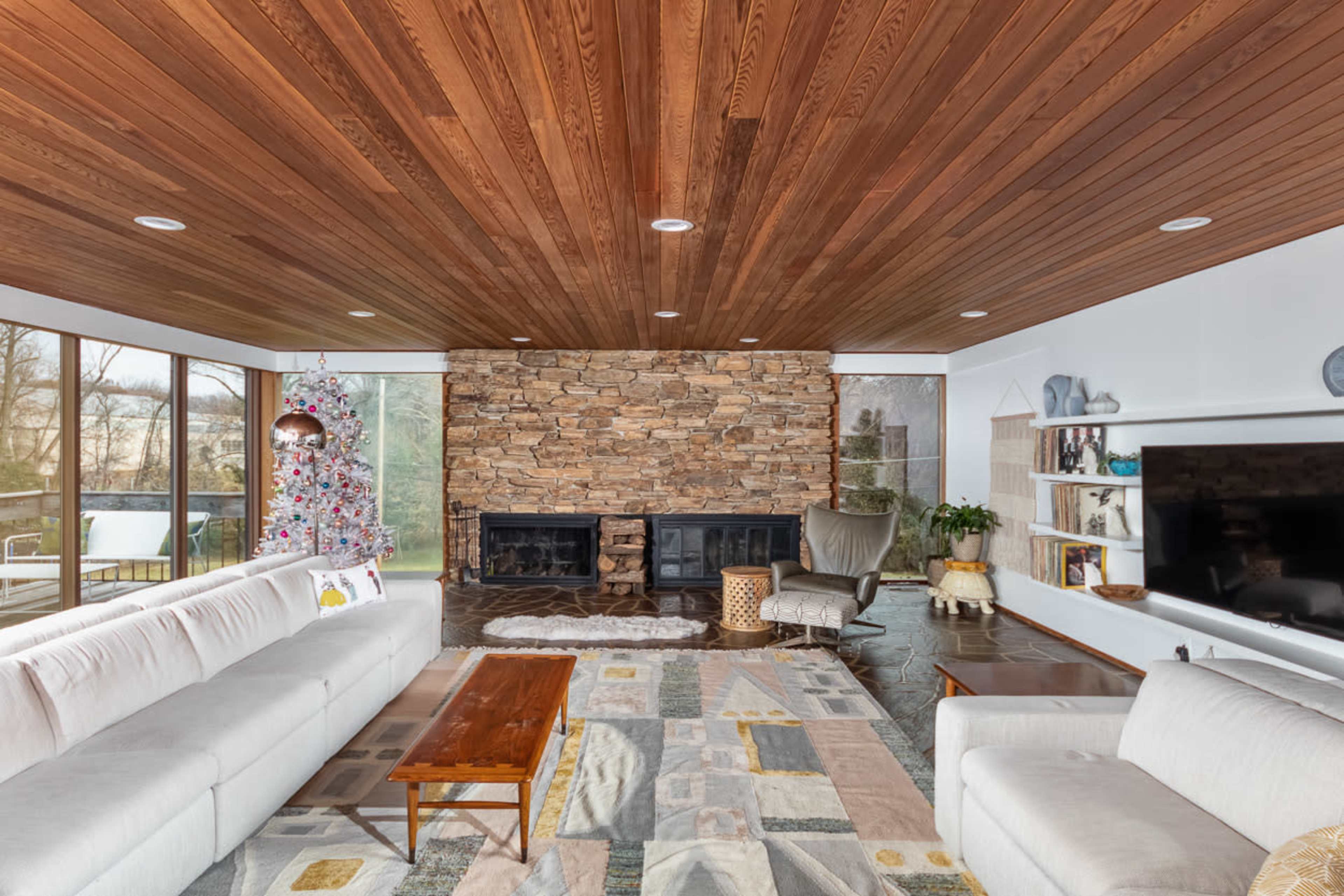 The living room features a wooden ceiling, stone-accented wall with two fireplaces, and large windows allowing natural light, complemented by minimalistic furniture and a decorative tree.