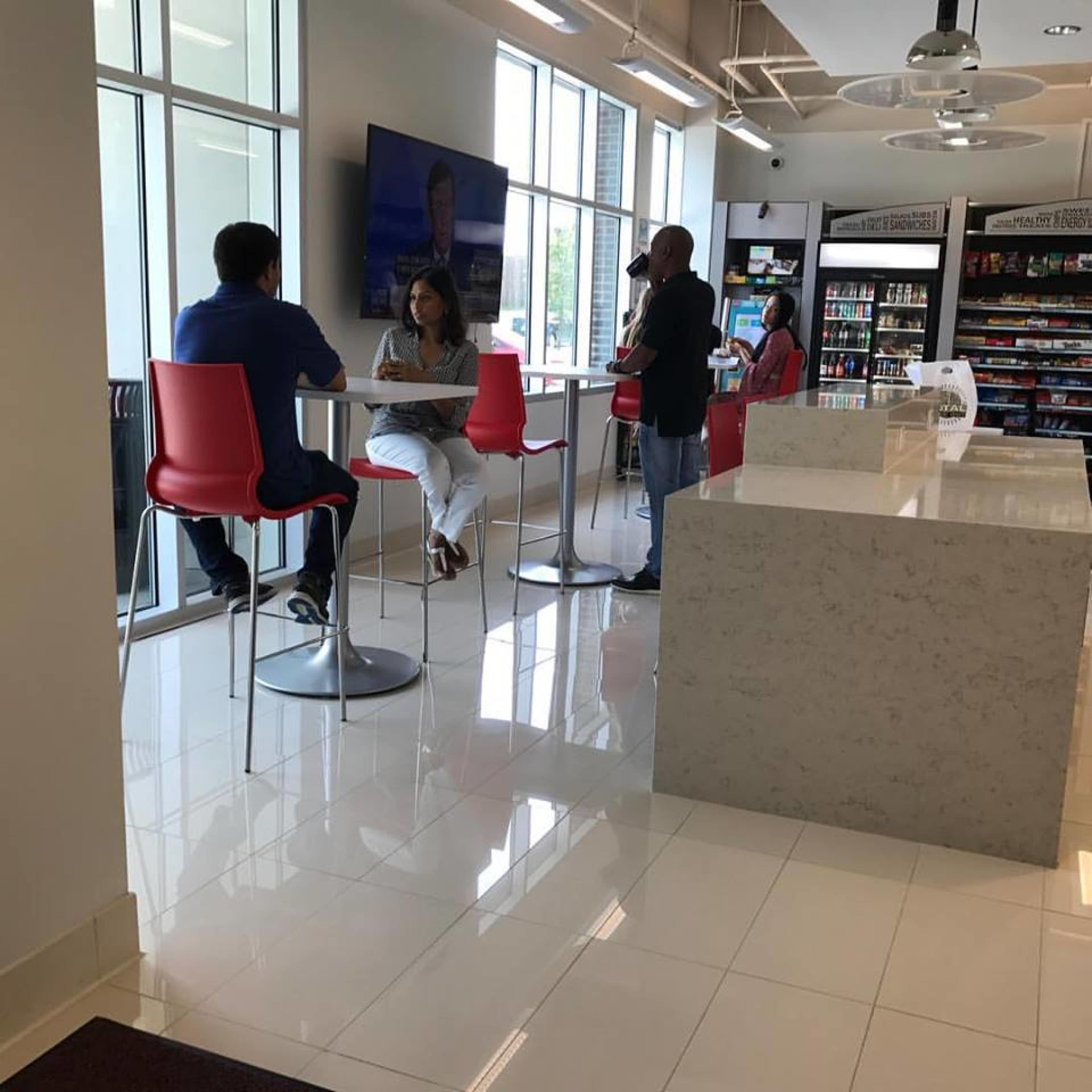 A modern café features a seating area with people interacting at high tables, alongside a counter displaying various snacks and beverages.