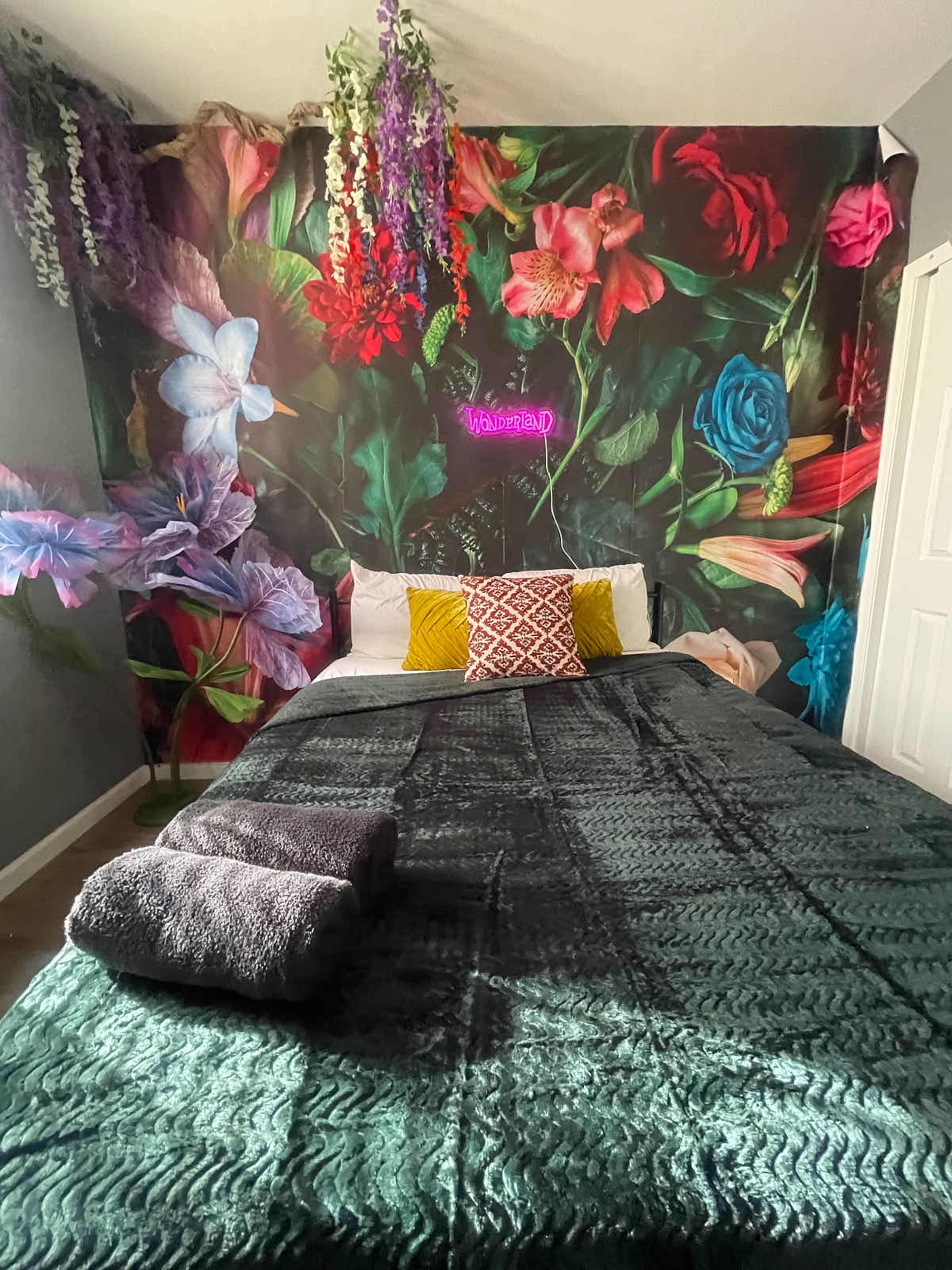 The image shows a bedroom with a dark floral feature wall, a bed with a green blanket, decorative pillows, and a neon sign.