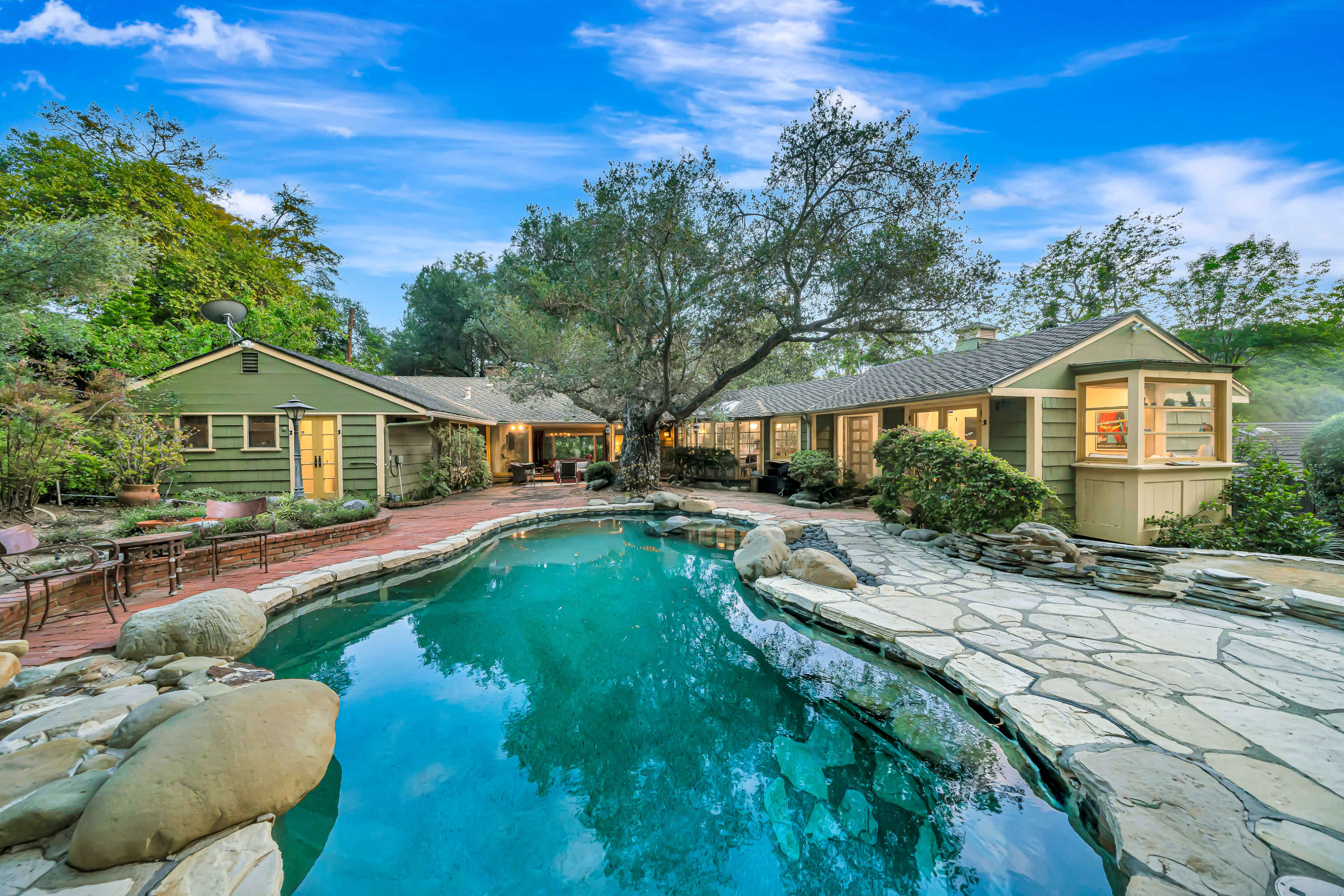 A residential property features a swimming pool surrounded by stone landscaping and a green-toned house with large windows set amidst lush trees.