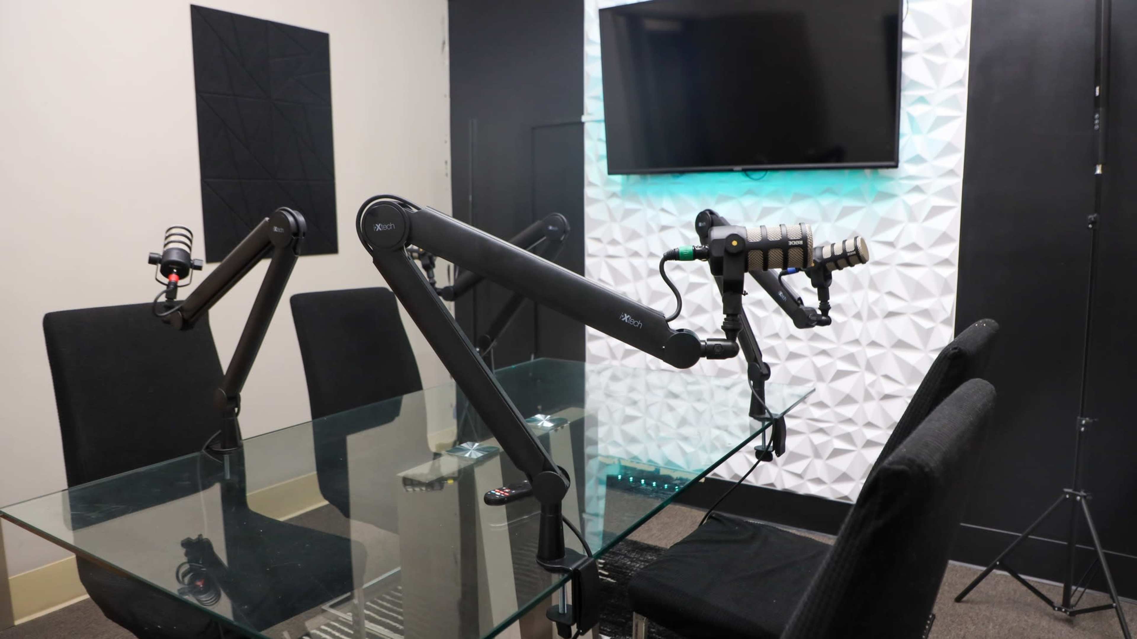 A modern recording studio setup featuring two boom microphone arms positioned over a glass table with black chairs, and a television mounted on a textured white wall in the background.