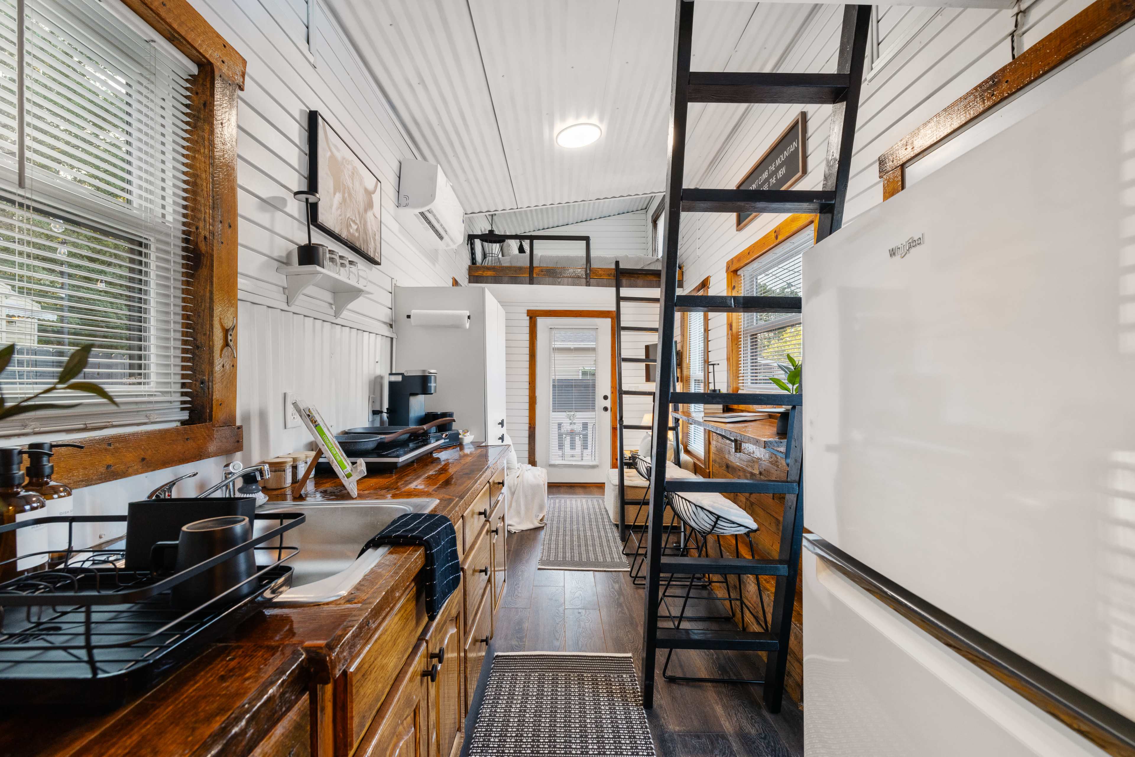 The image shows a small, well-organized kitchen area in a tiny home, featuring a wooden countertop, a sink, a refrigerator, and a staircase leading to a loft.