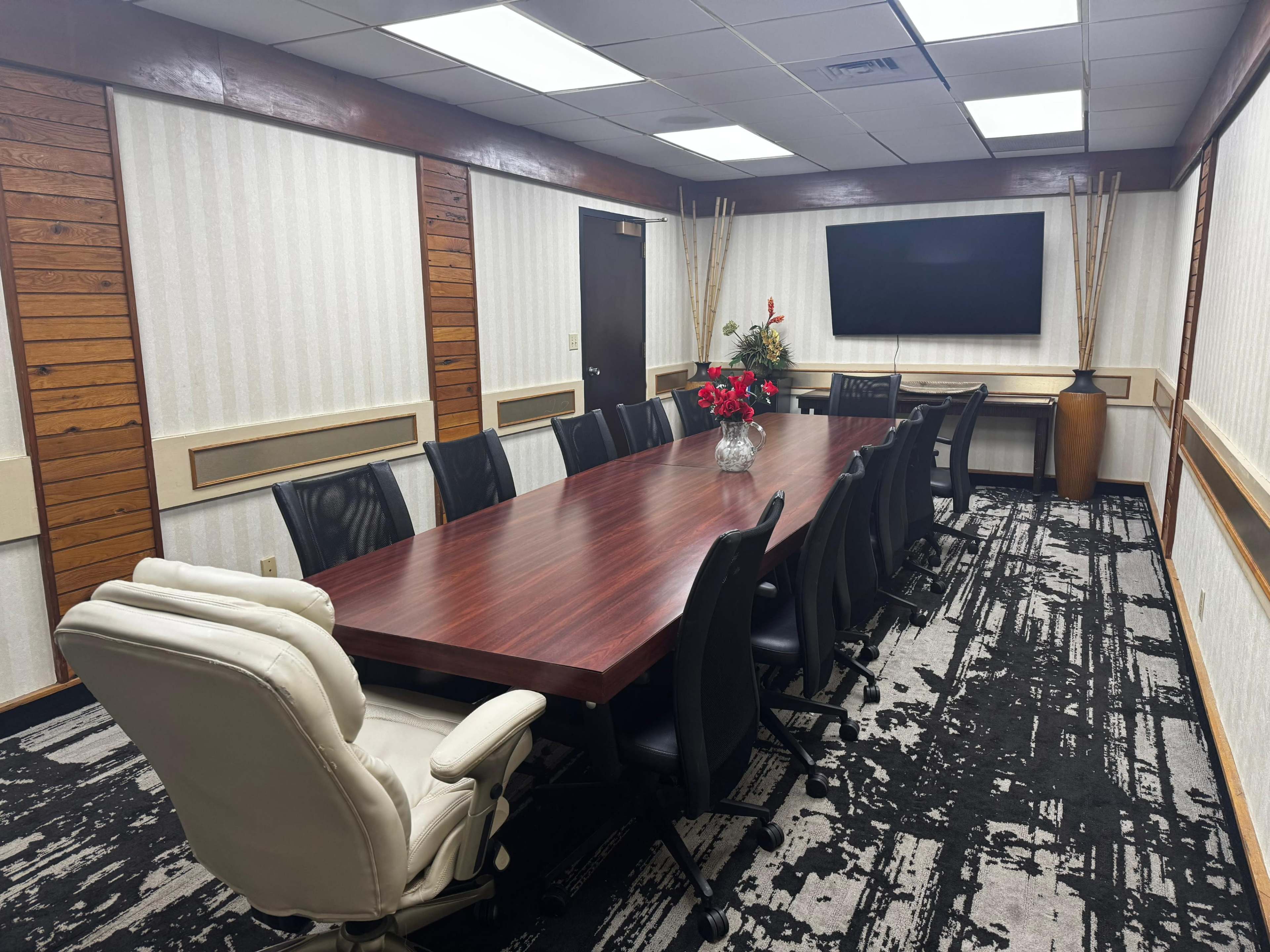 The image shows a conference room with a large wooden table surrounded by black office chairs, a white executive chair, a television mounted on the wall, and decorative plants.