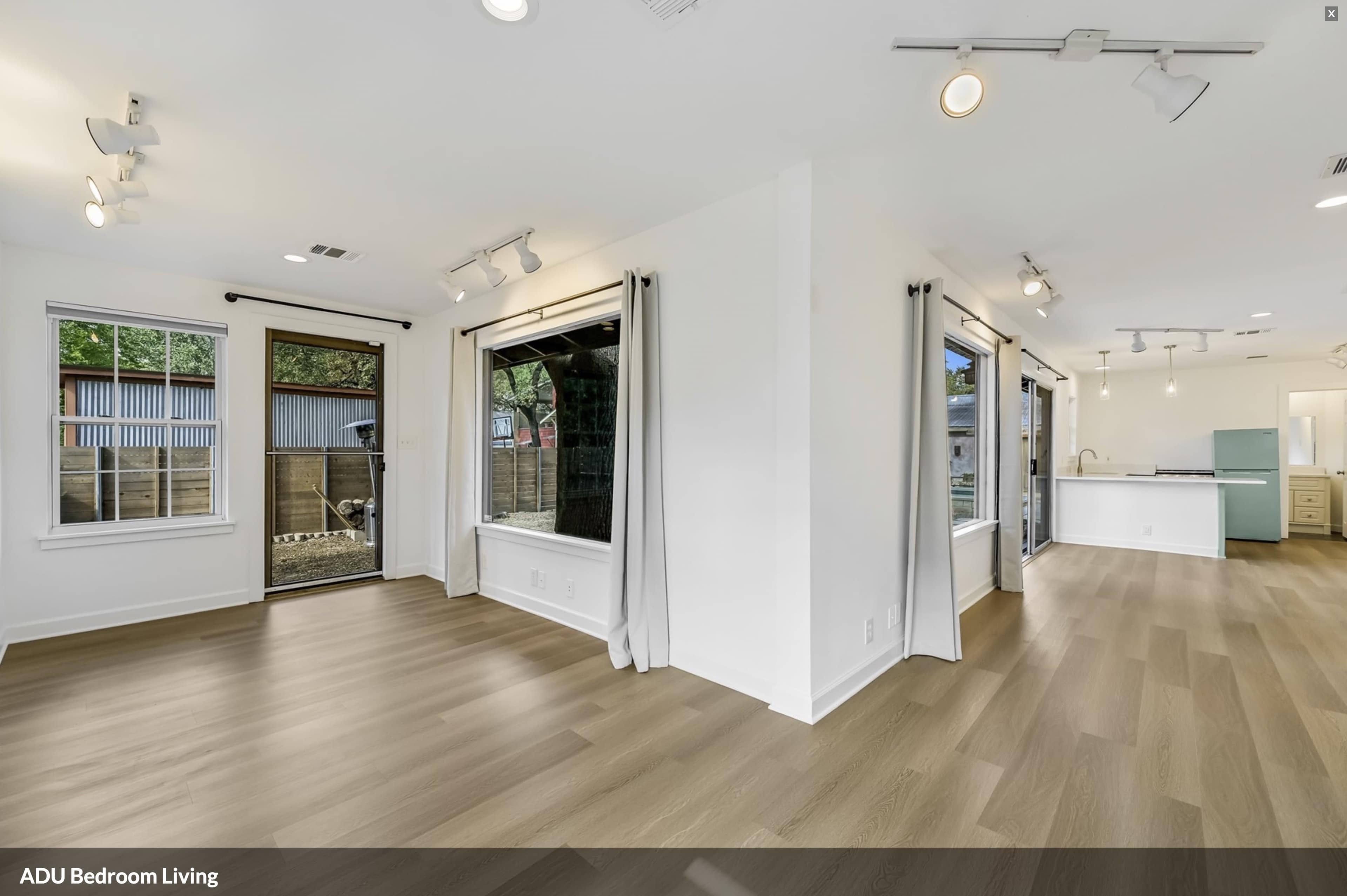 The image shows a spacious, empty room featuring large windows, hardwood flooring, and an open layout with visible access to a kitchen area.