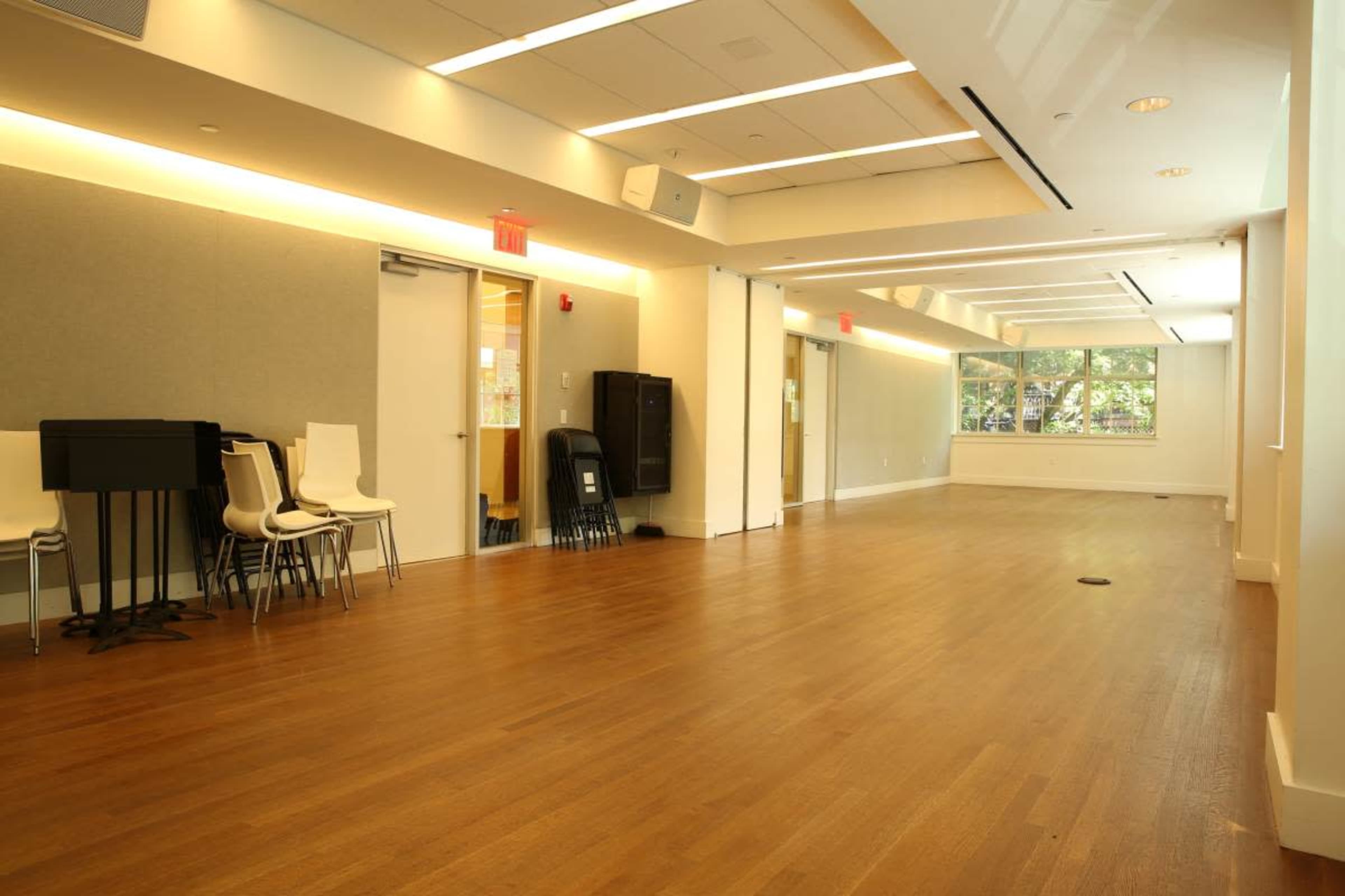 The image shows a large, empty room with wooden flooring, bright lighting, and several chairs stacked against the wall.