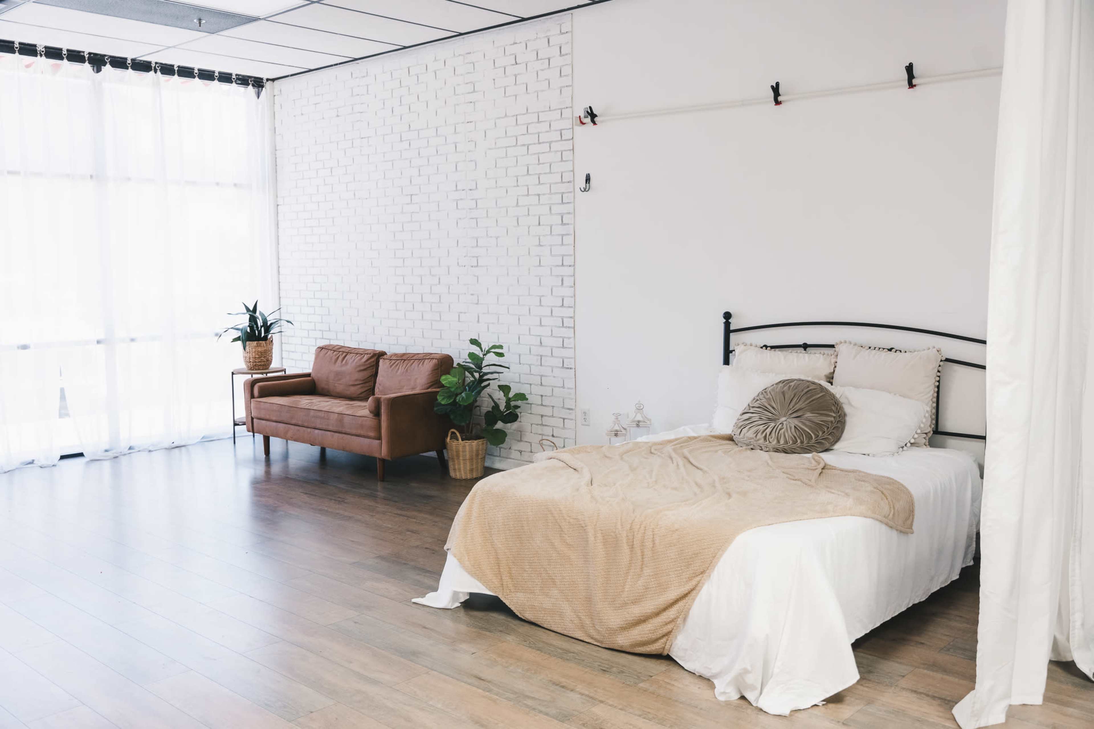 A bedroom features a bed with a beige blanket and a brown couch against a white brick wall, complemented by potted plants and large windows with sheer curtains.