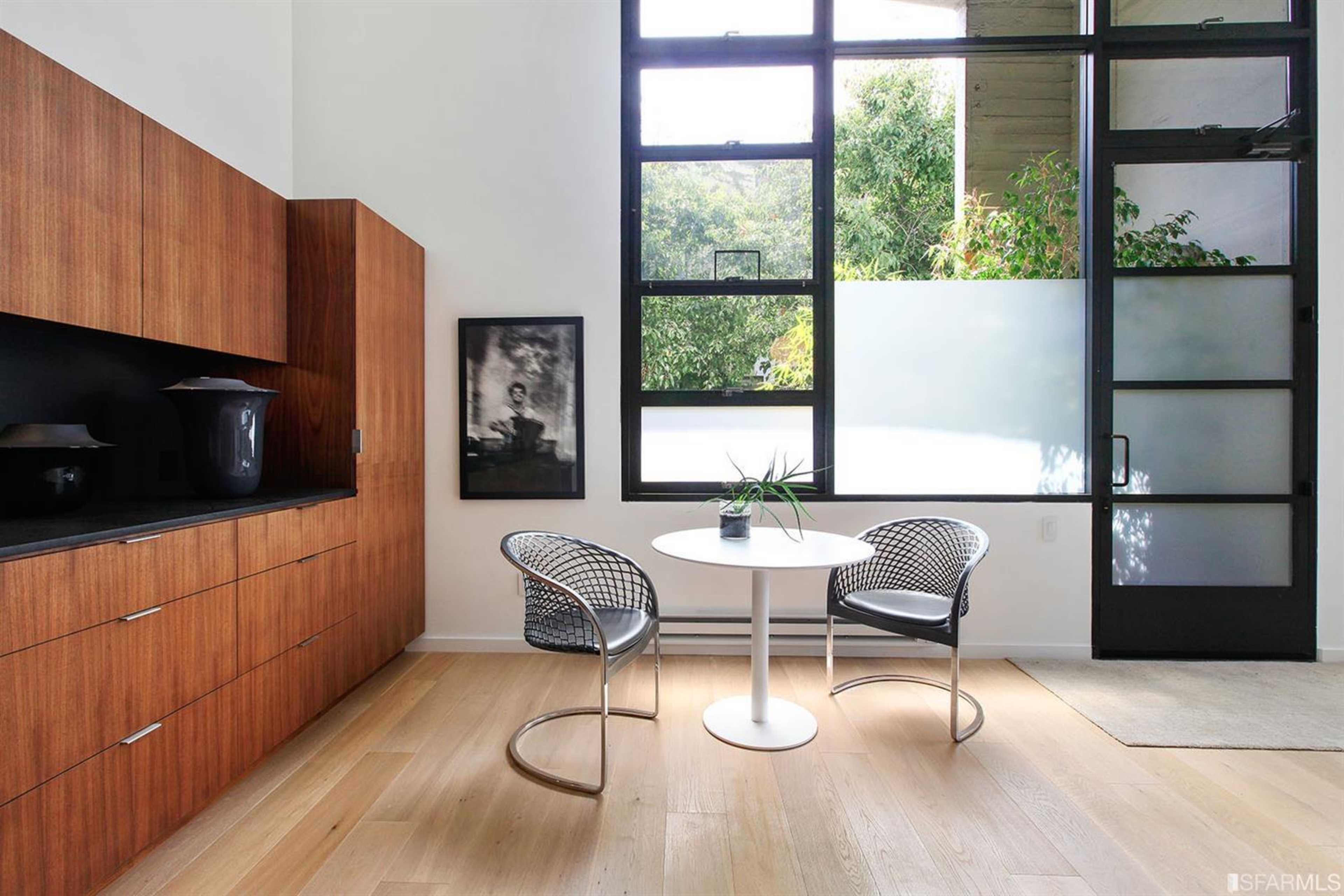 A modern interior with a round white table and two wire chairs beside a large window that offers a view of greenery.