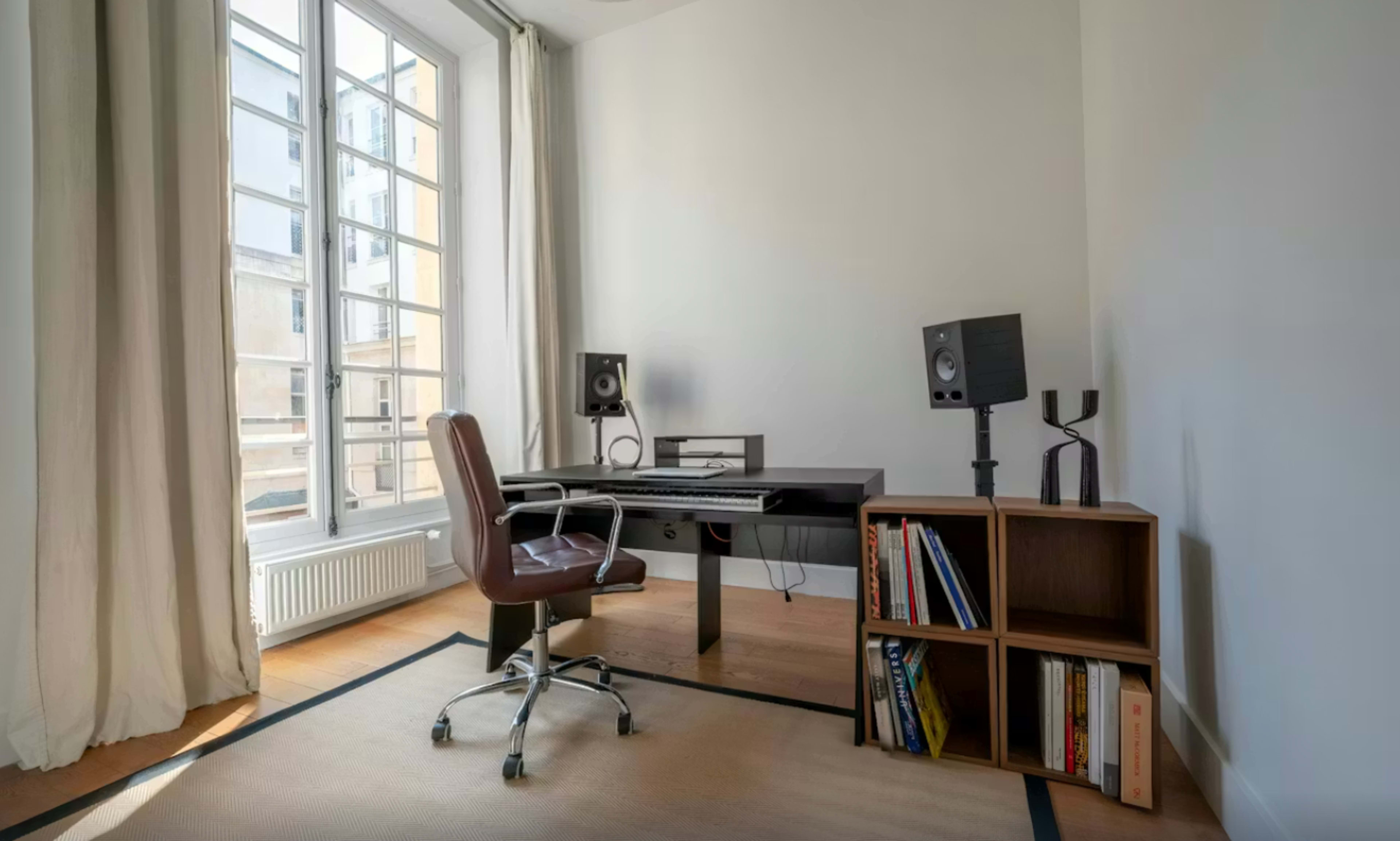 The image shows a modern home office featuring a black desk with an ergonomic brown chair, two studio monitors on stands, and a wooden bookshelf containing books.
