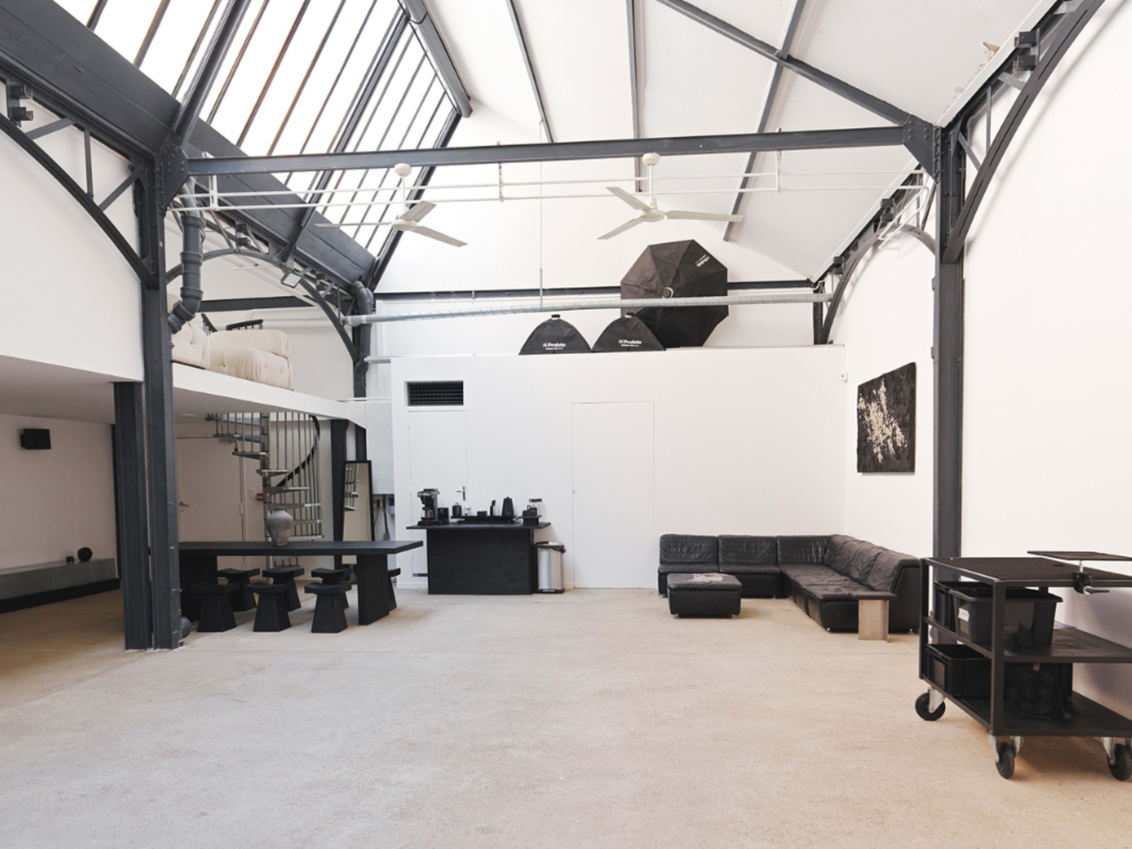 The image shows a spacious, modern studio with high ceilings, large windows, and minimalist furniture including a black sofa and a table.