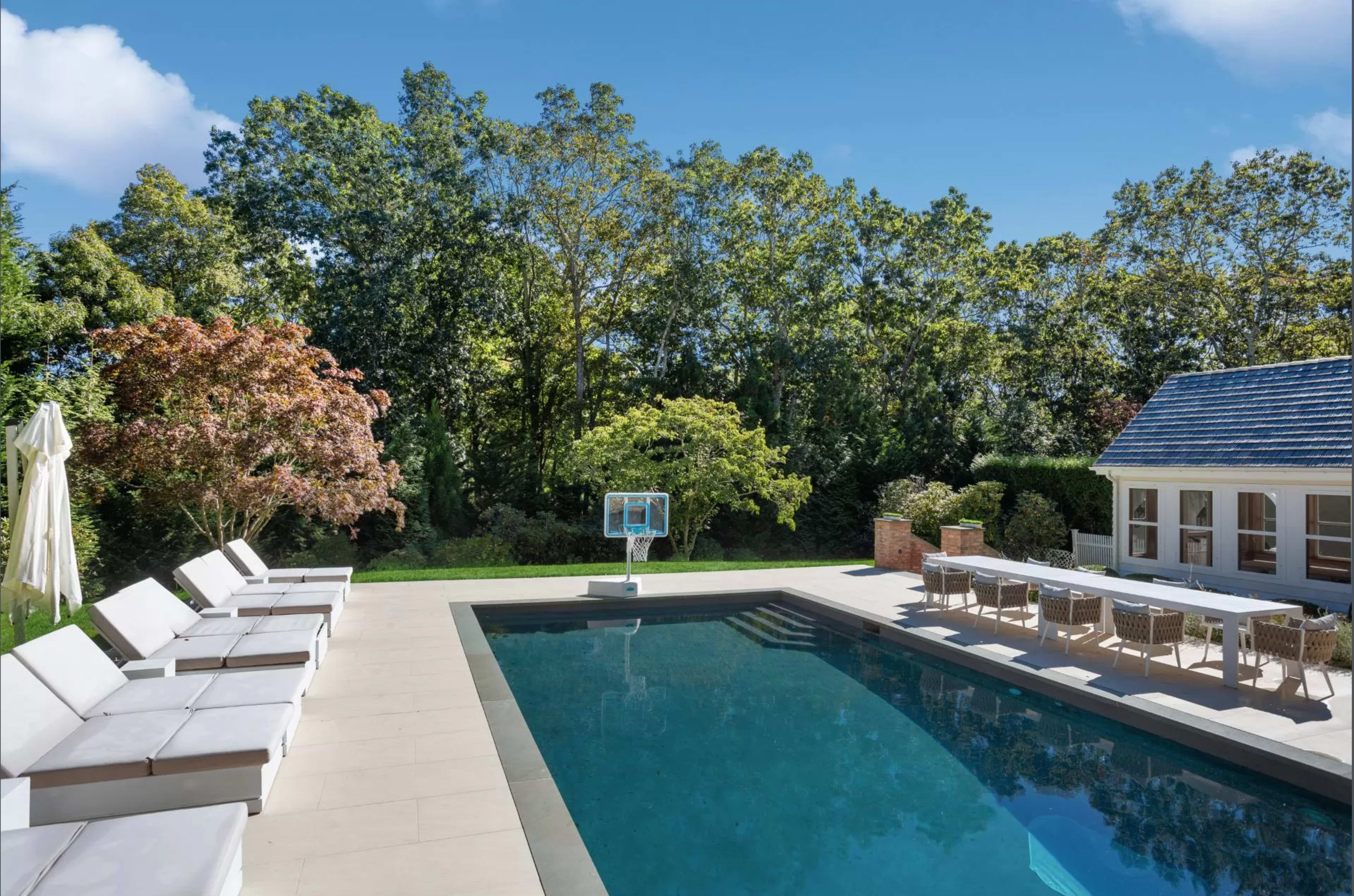 A modern swimming pool is surrounded by lounge chairs and backed by lush greenery and a house with large windows.