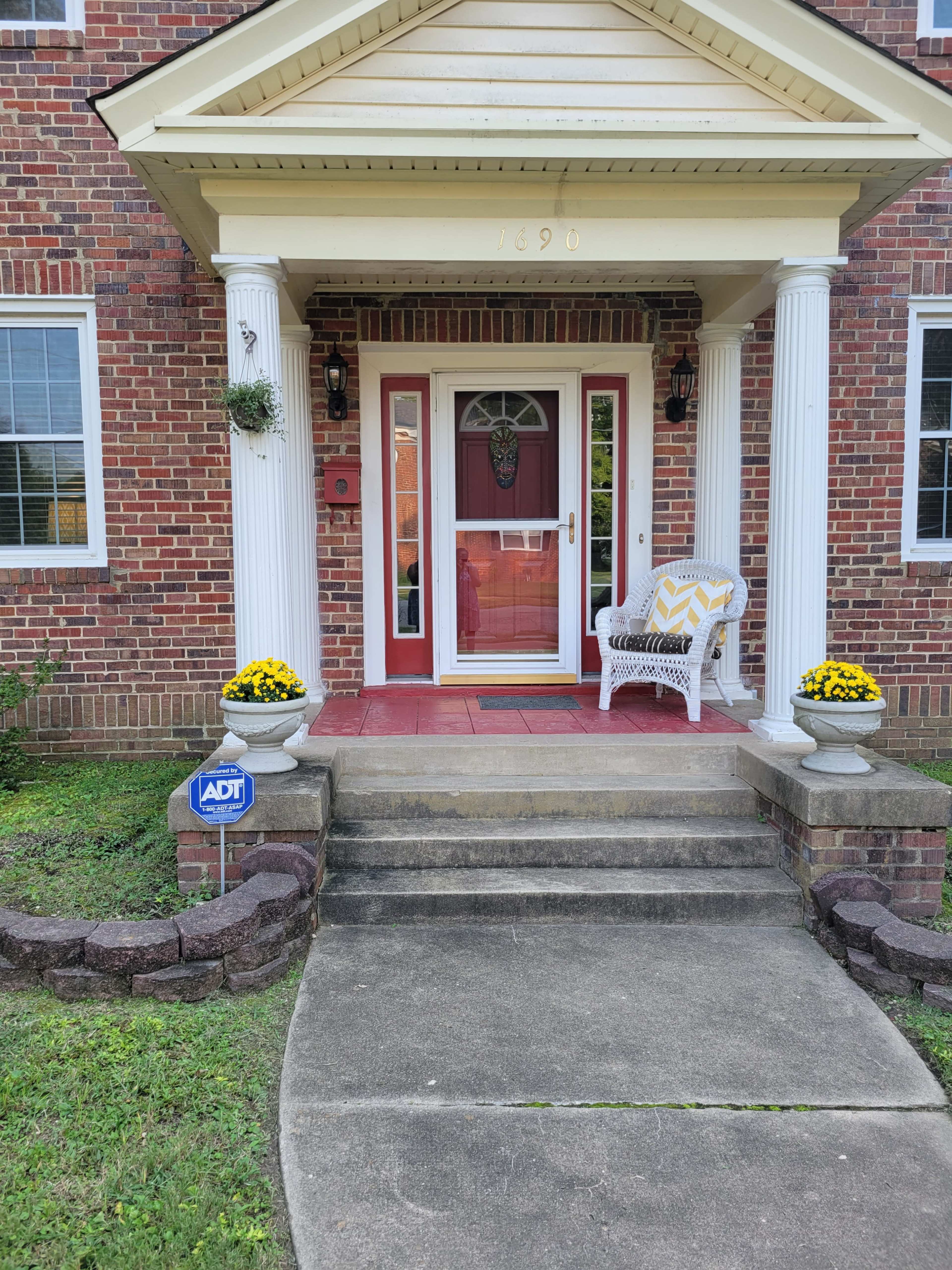 The image shows a front entrance of a house with brick siding, featuring a red door, white columns, and potted yellow flowers on either side of the steps.