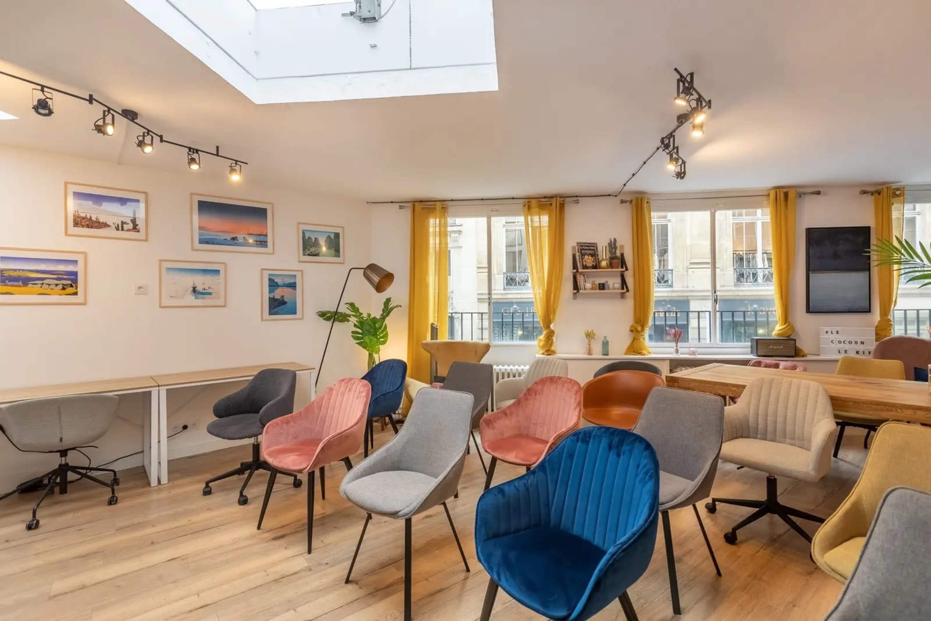 The image shows a bright, airy room with various colorful chairs arranged around a central space, featuring large windows with yellow curtains and framed pictures on the walls.