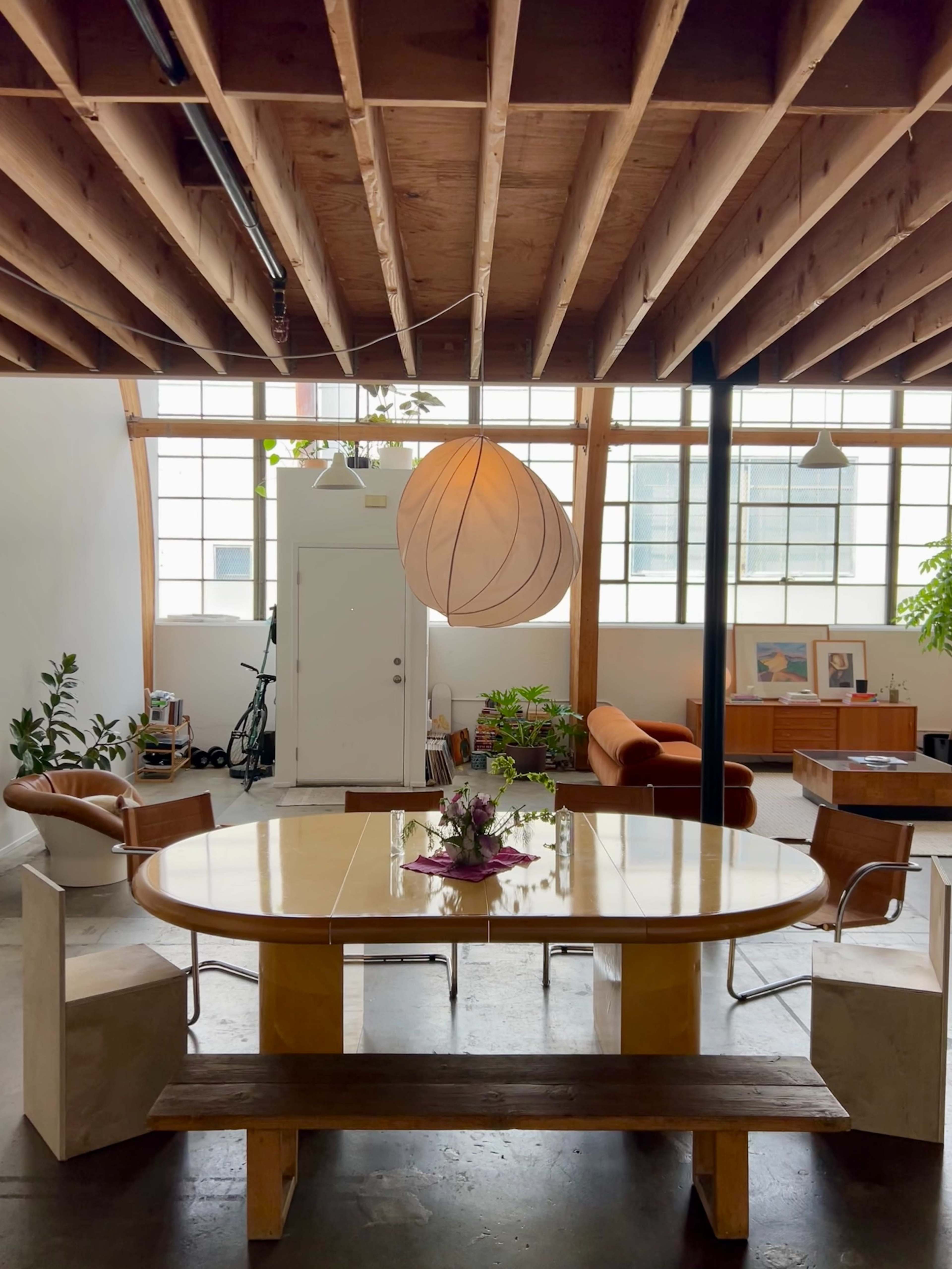 A spacious indoor area featuring a round dining table surrounded by chairs, a large pendant light, and a mixture of plants and furniture against a backdrop of large windows.