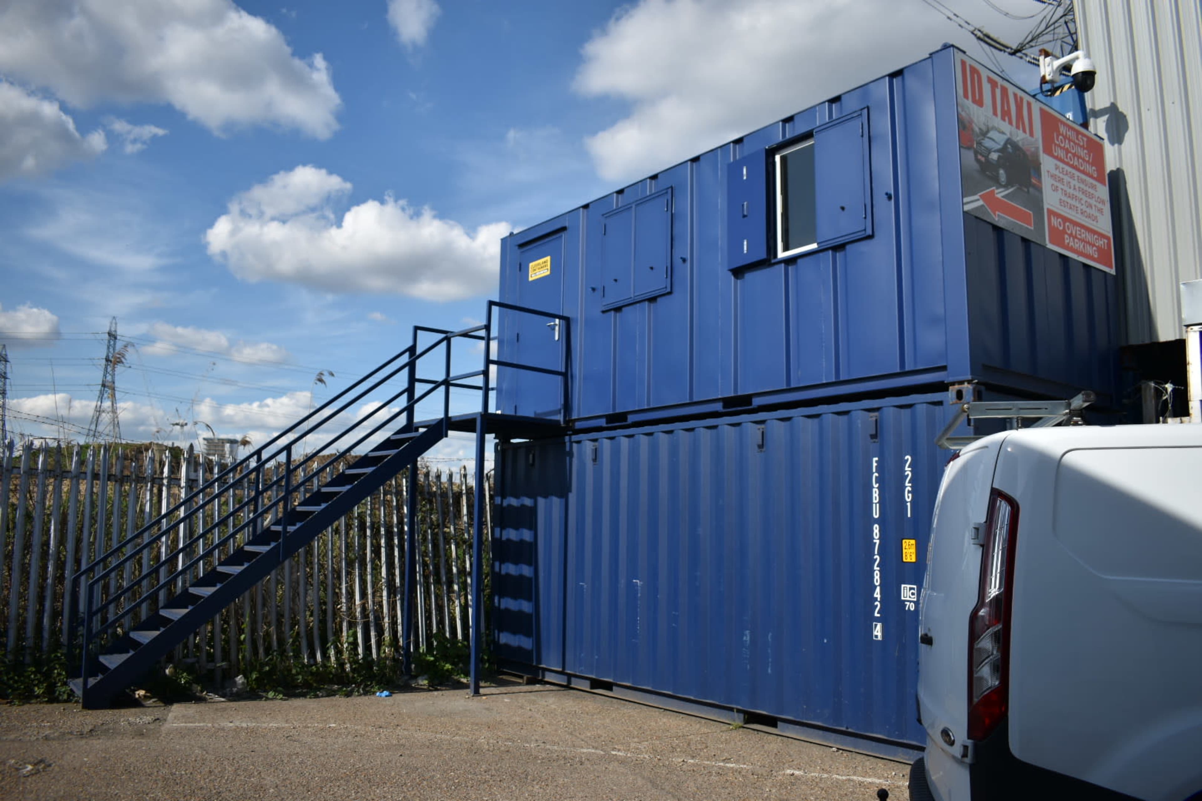 A blue shipping container is stacked on top of another, with a staircase leading to a door, and a sign on the wall nearby.
