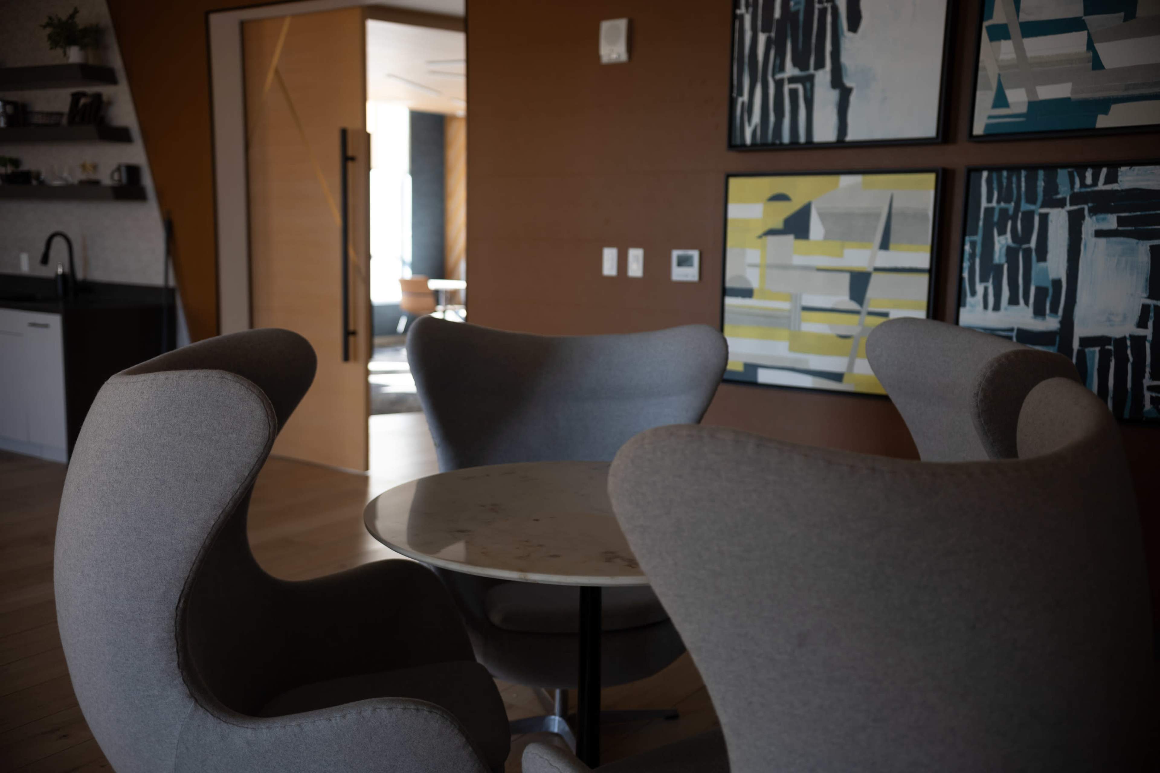 The image shows a modern interior space with four curved gray chairs surrounding a circular table and abstract art on the walls.