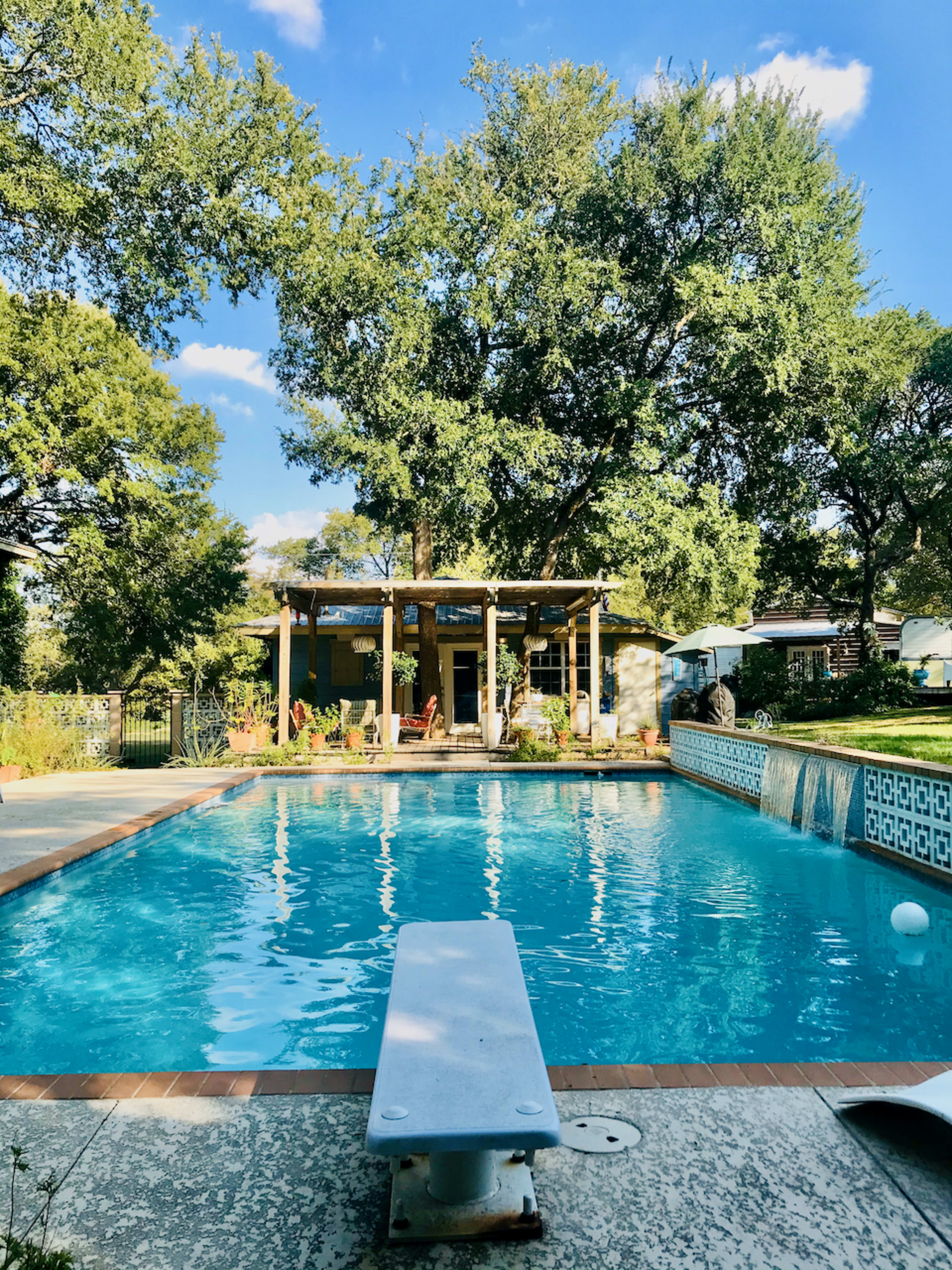 A clear swimming pool features a diving board and is surrounded by trees and a house with a shaded patio in the background.