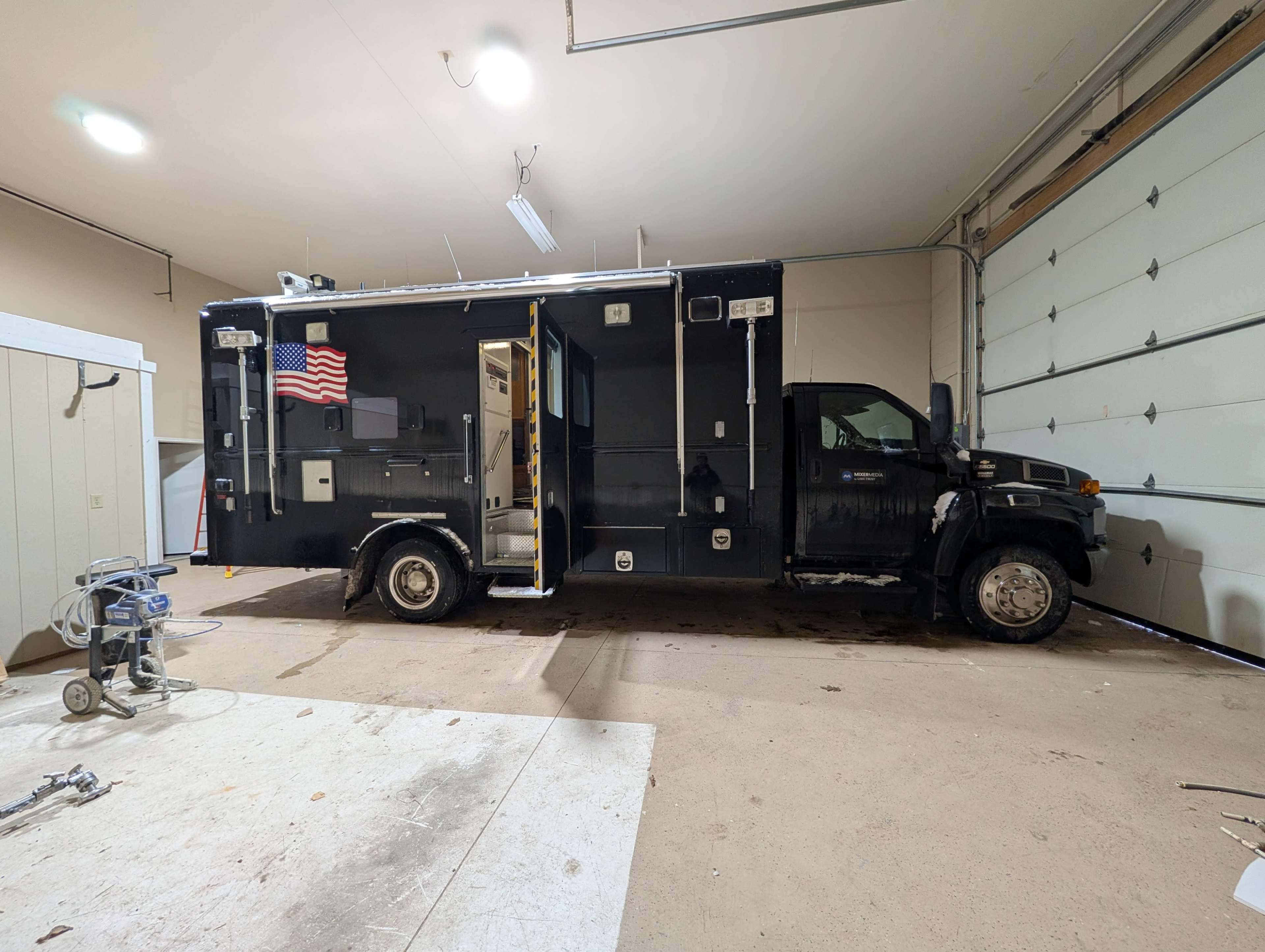 A large black mobile command unit is parked inside a spacious garage, with its side door open and an American flag displayed.