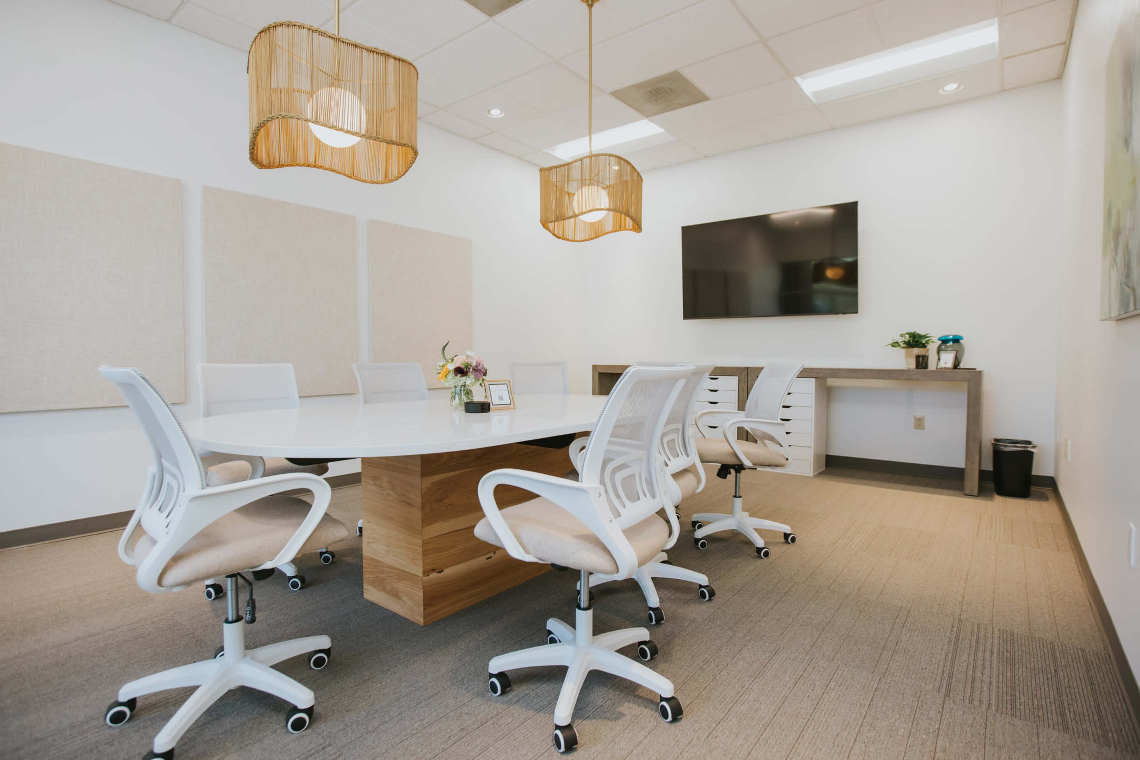 A modern conference room features a large white table surrounded by ergonomic chairs, with soft lighting and a wall-mounted television.