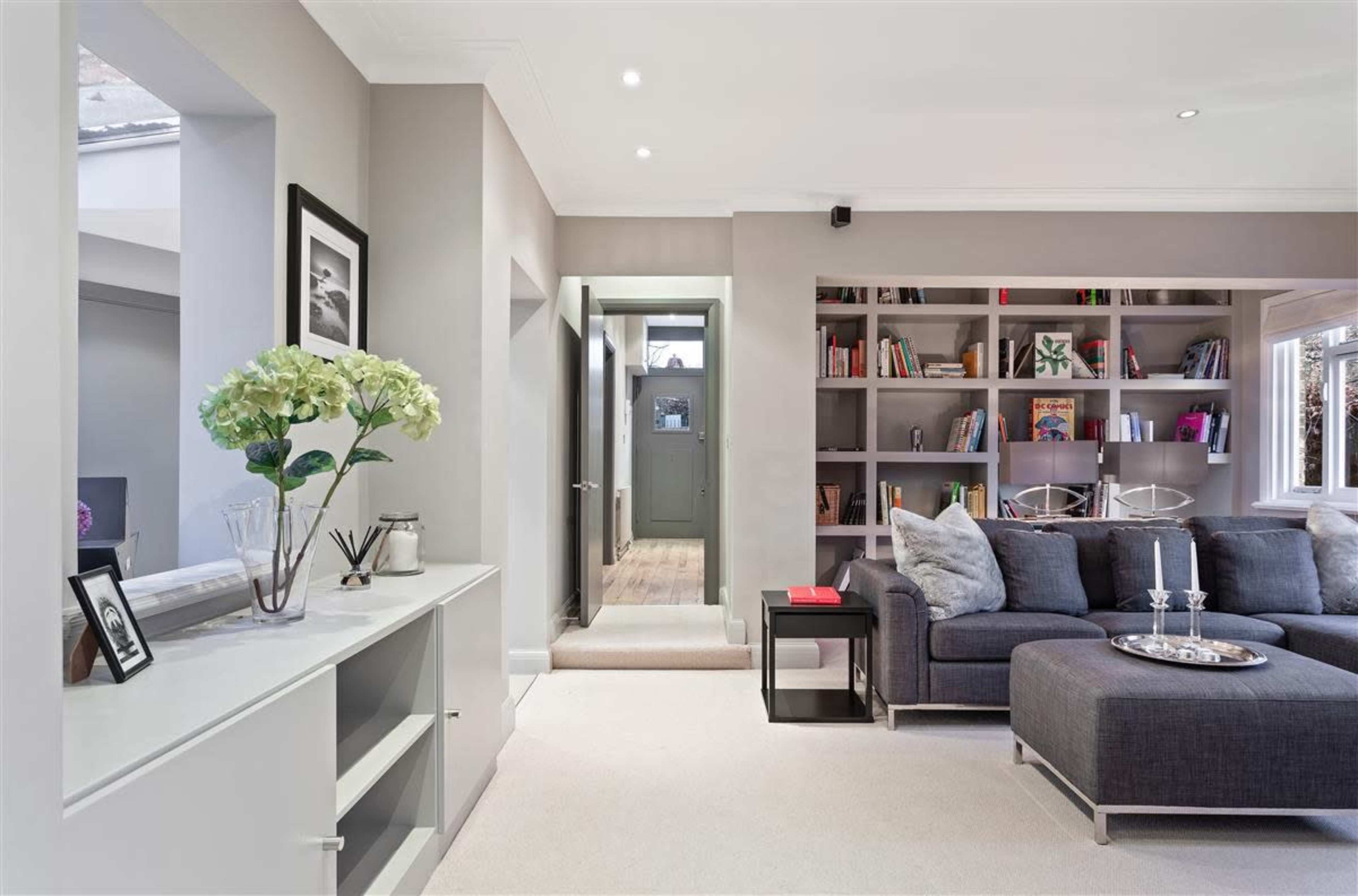 A modern living room featuring a gray sectional sofa, a bookshelf filled with books and decor, and a hallway leading to a door.