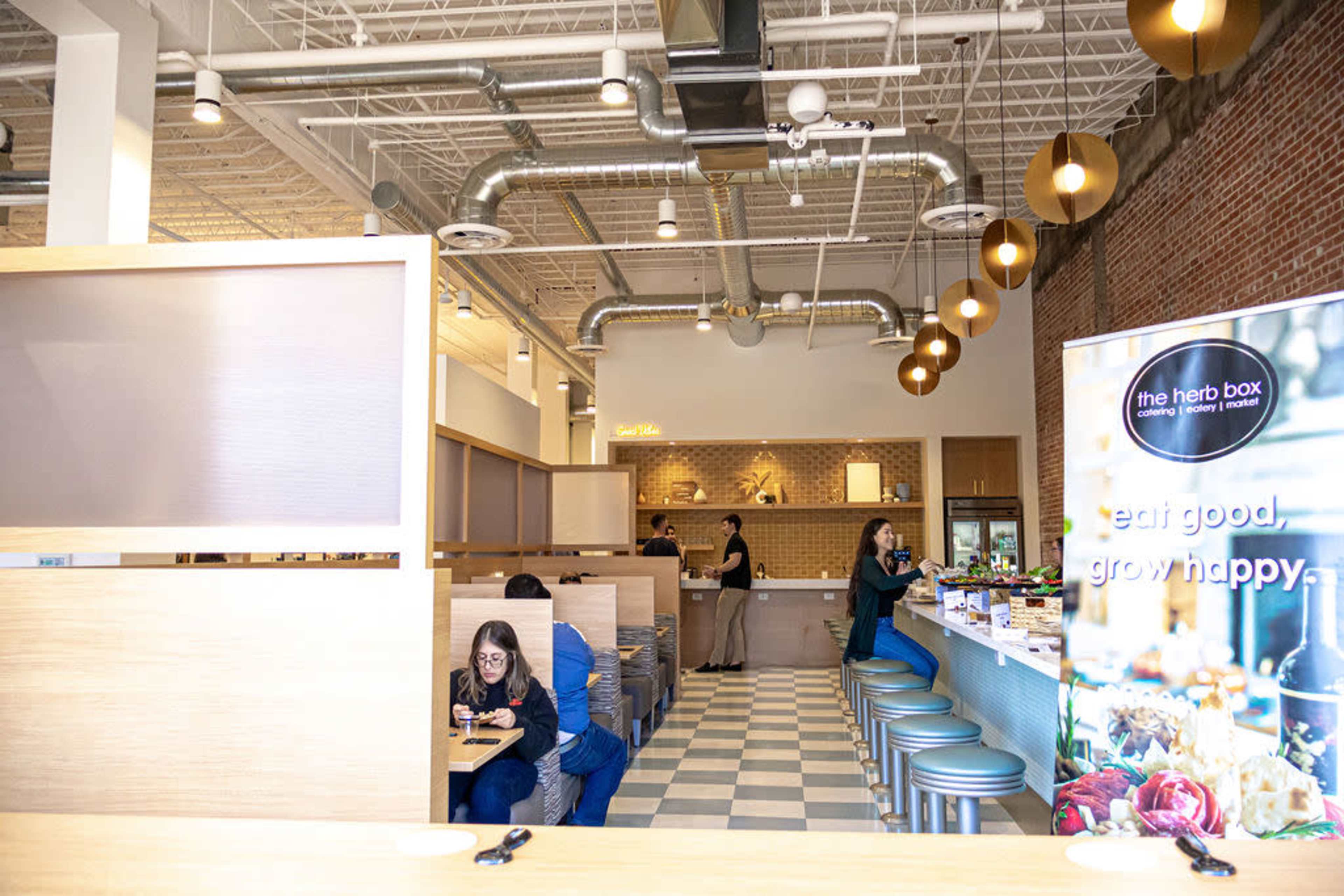 The image shows a modern dining area of a restaurant with wood and tile decor, where patrons are seated at tables and a staff member is preparing food behind the counter.