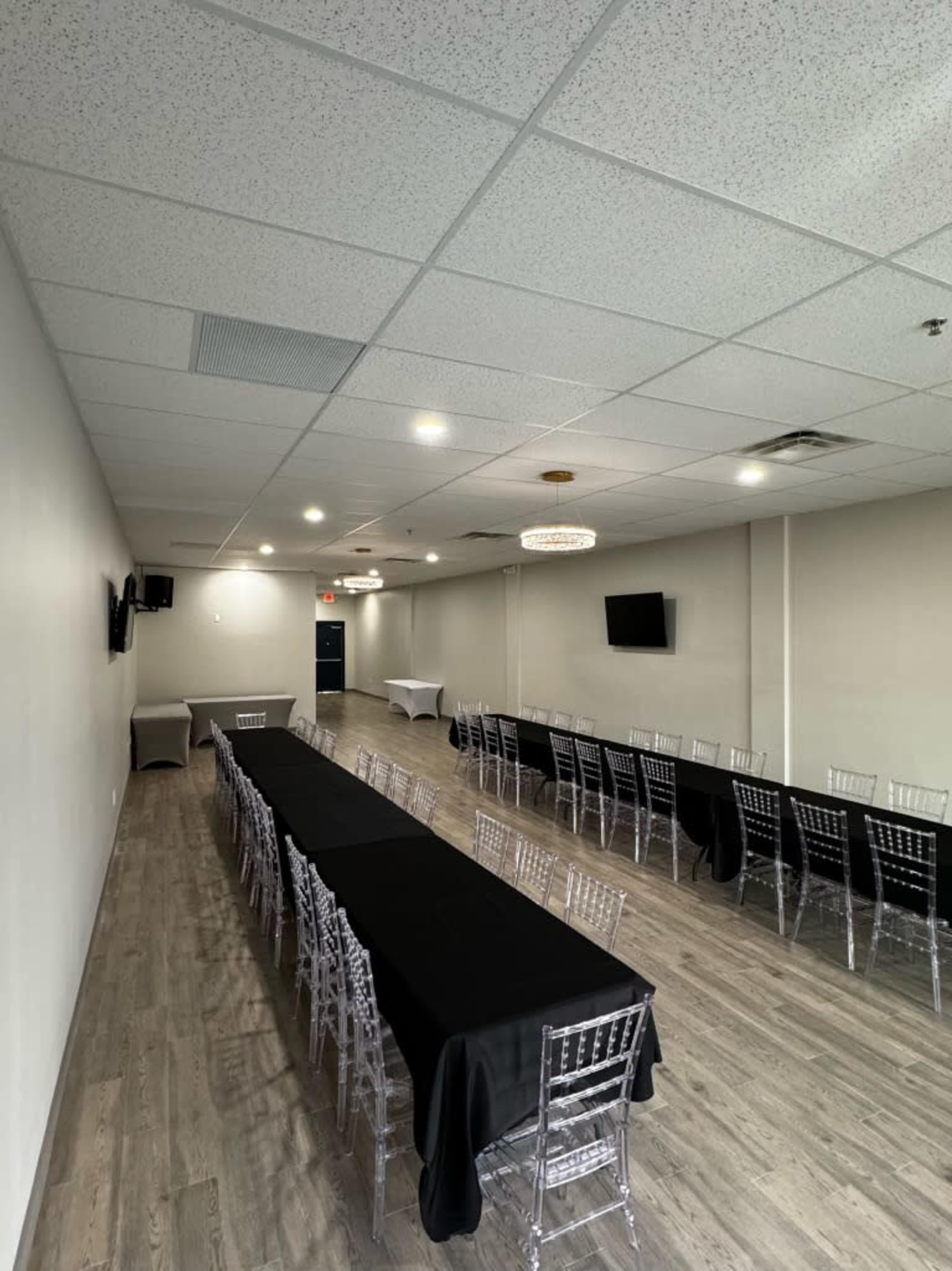 A spacious meeting room features long tables covered with black tablecloths and clear plastic chairs arranged in rows, with modern lighting overhead.