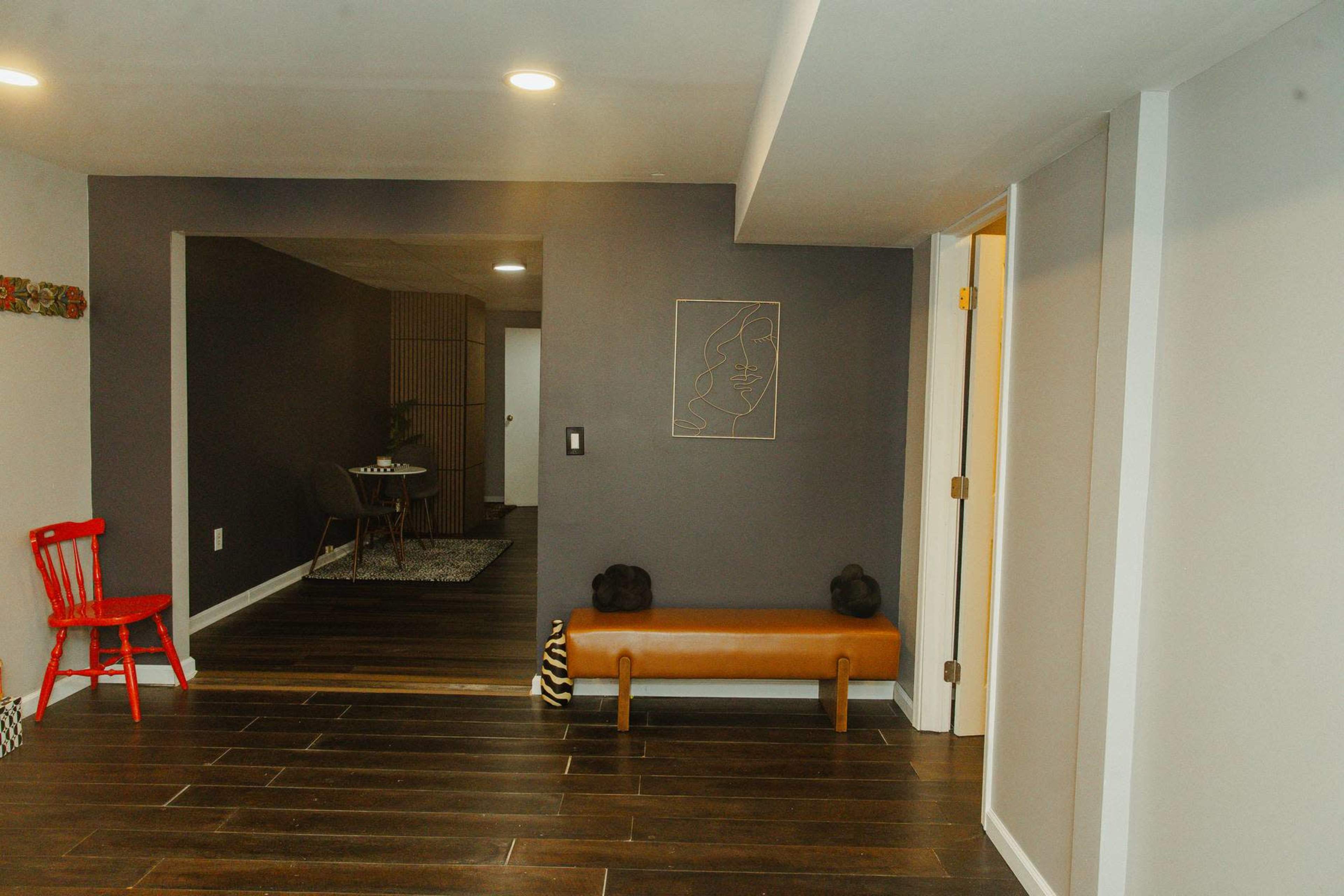 The image shows a hallway with a wooden bench, a red chair, and a minimalist line art piece on the wall, leading to another room.