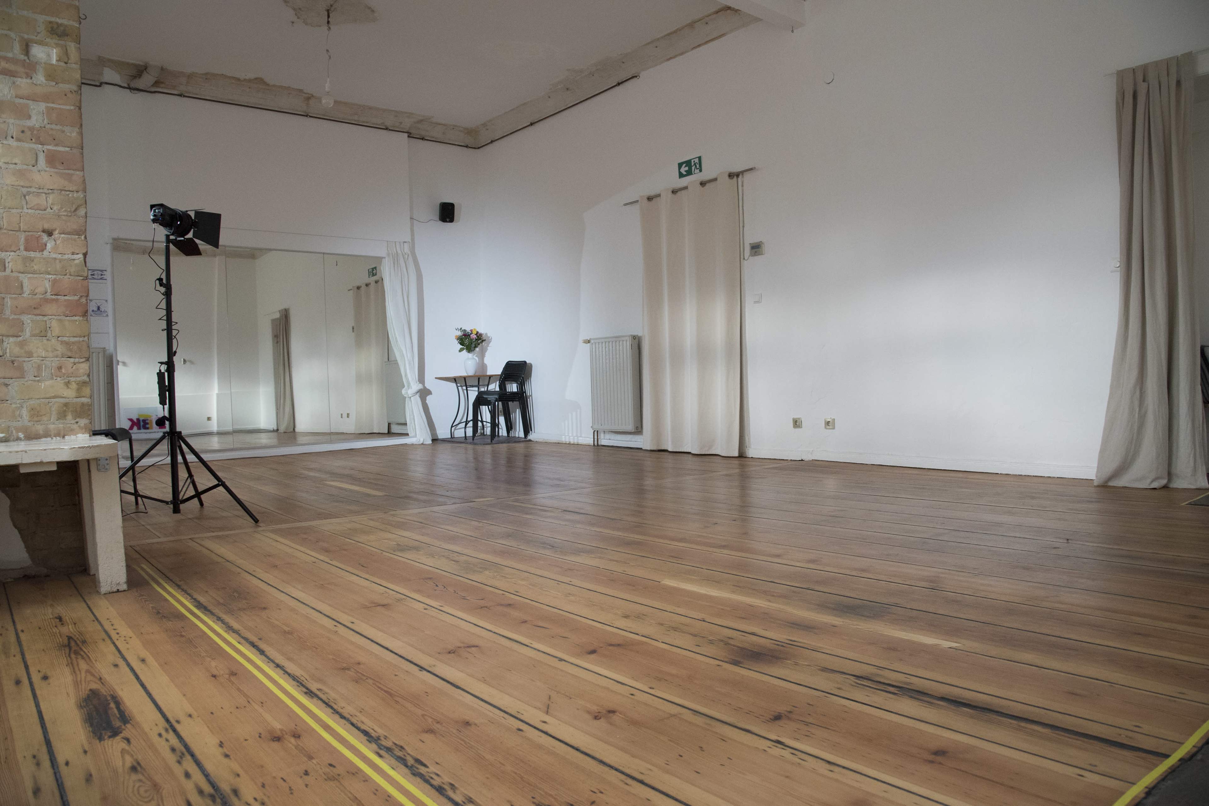 A spacious, empty room with wooden flooring, a standing light equipment, a chair, and partially closed curtains.