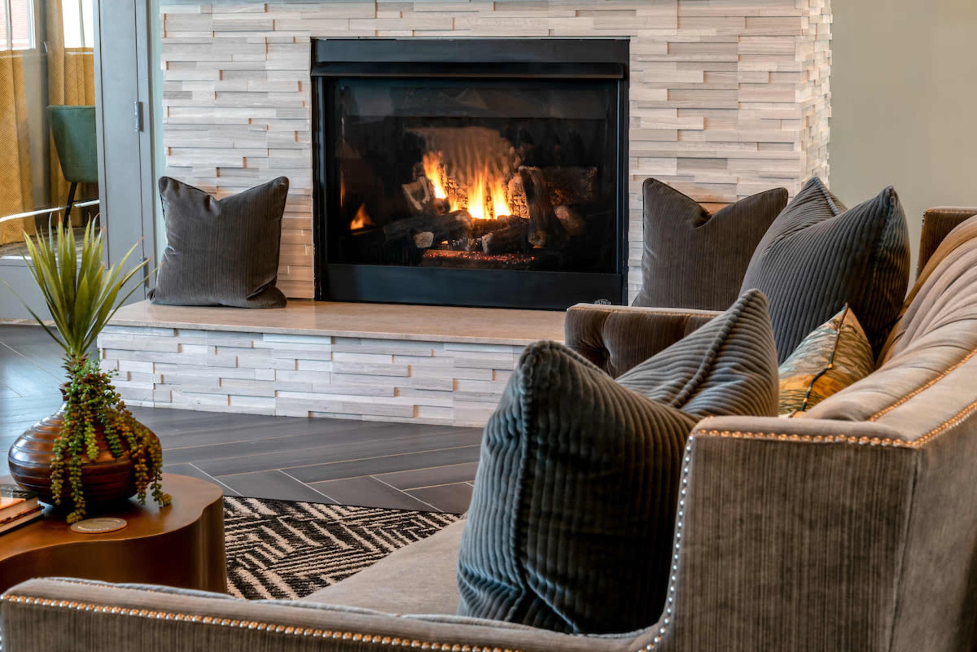 A modern living room features a stone fireplace with a lit fire, a comfortable sofa with decorative cushions, and a small table beside a plant.